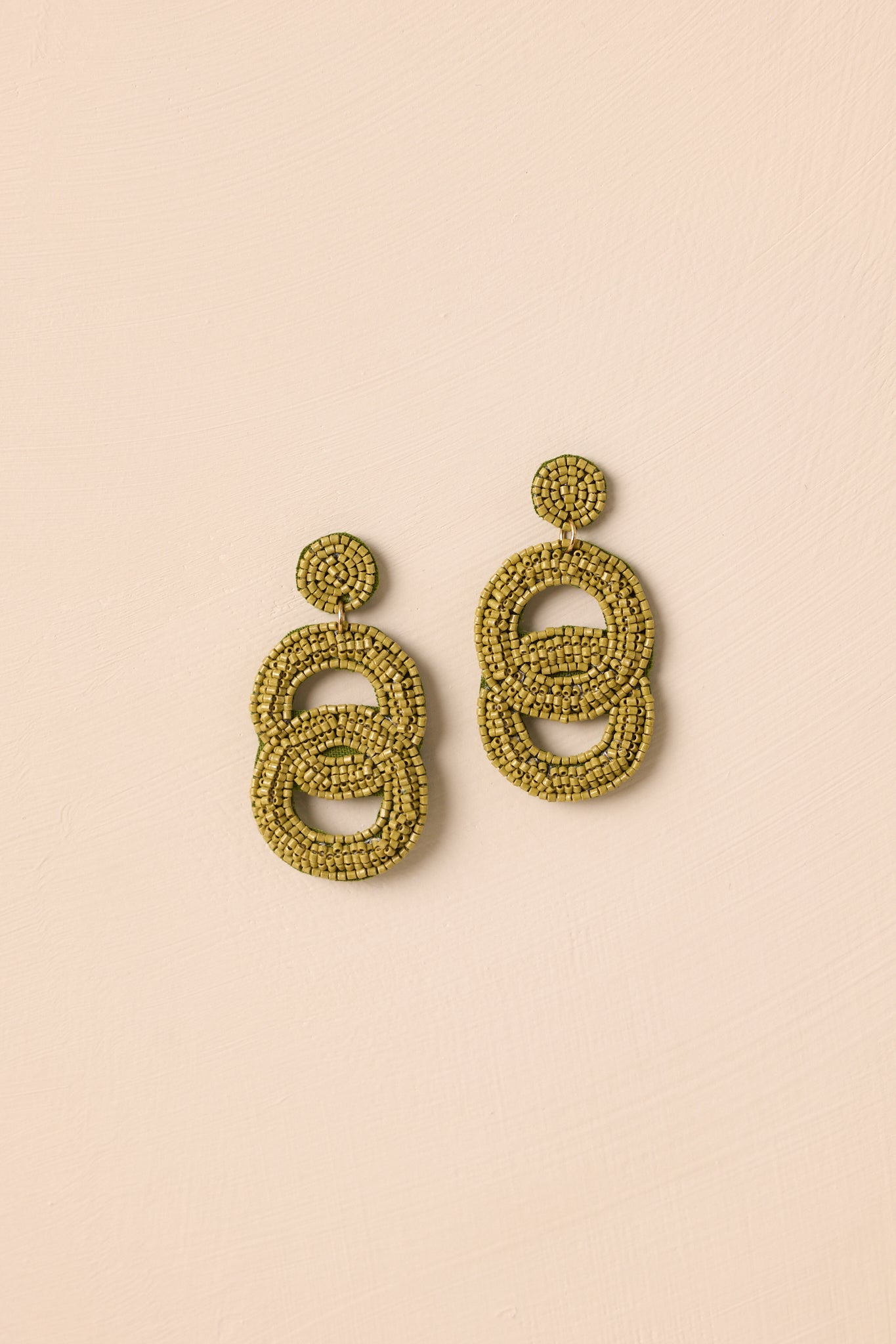 The earrings displayed against a beige background highlighting the olive green color and beaded texture.