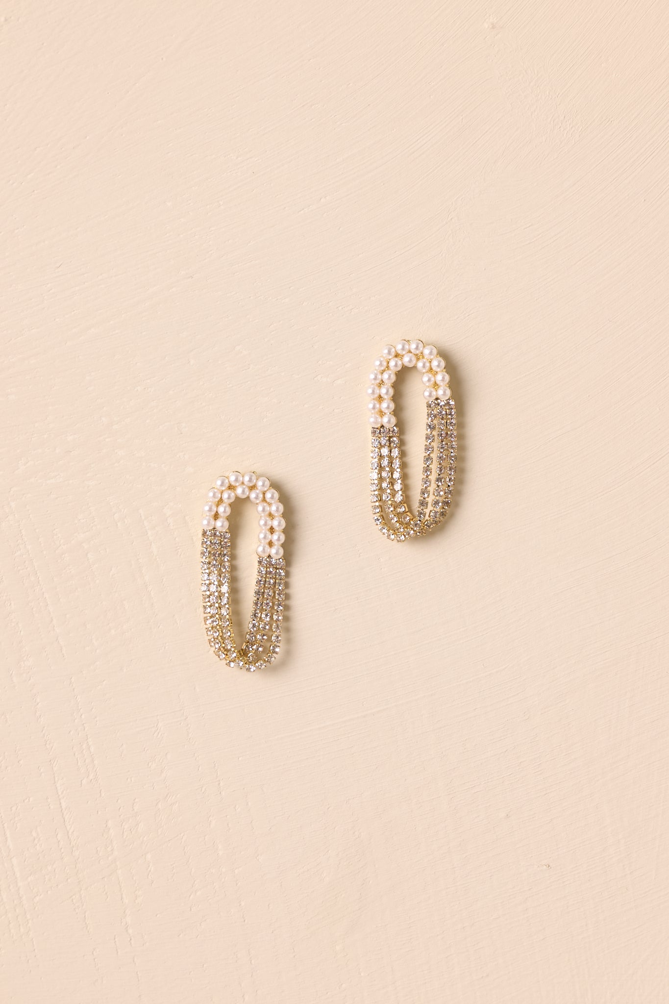 Elongated oval loop earrings displayed against a beige background, showcasing the layered chains with pearl and rhinestone accents.