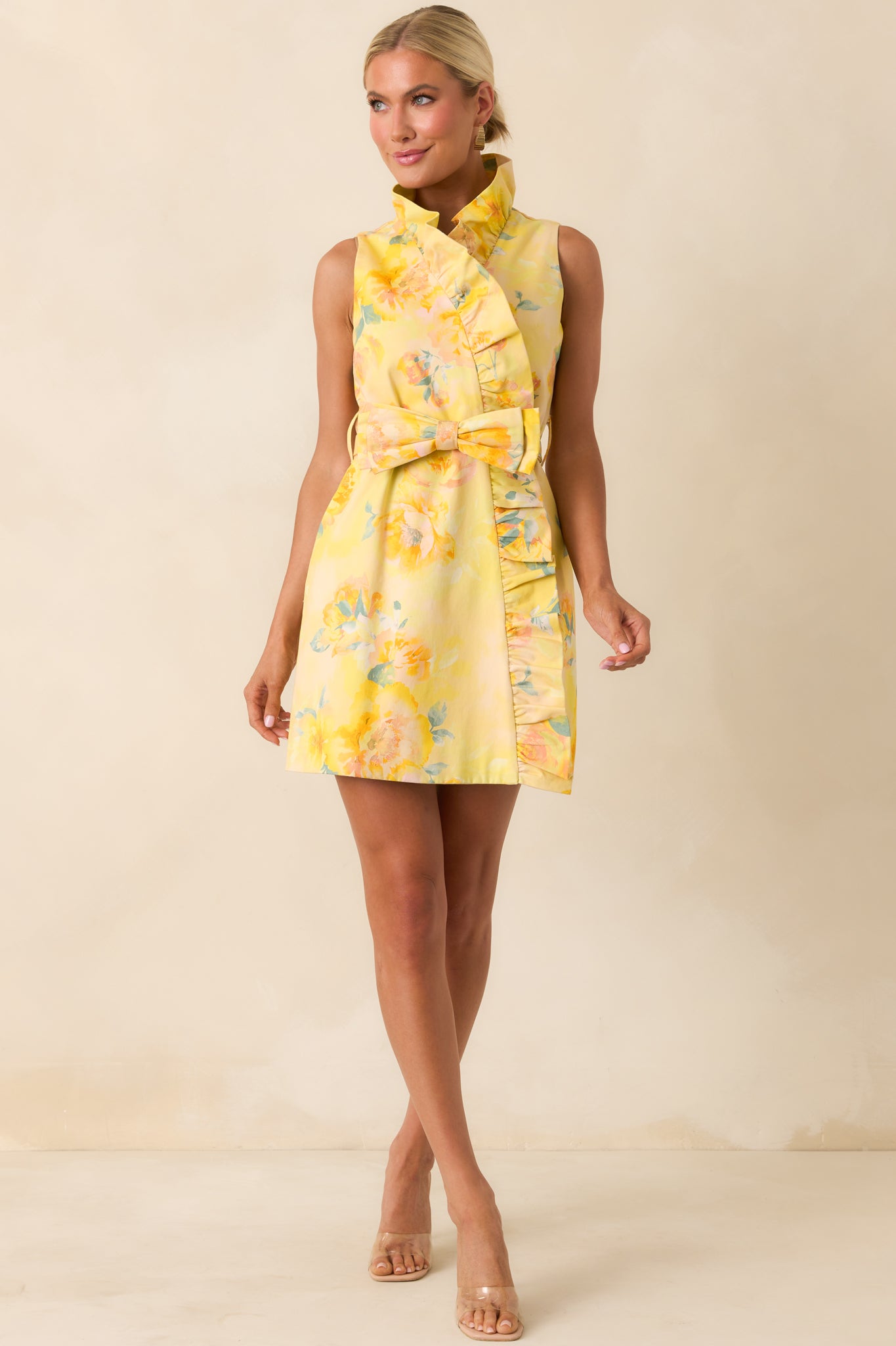 Floral mini dress in yellow tones featuring a soft ruffle neckline, self-tie waist belt, and wrap-style skirt.