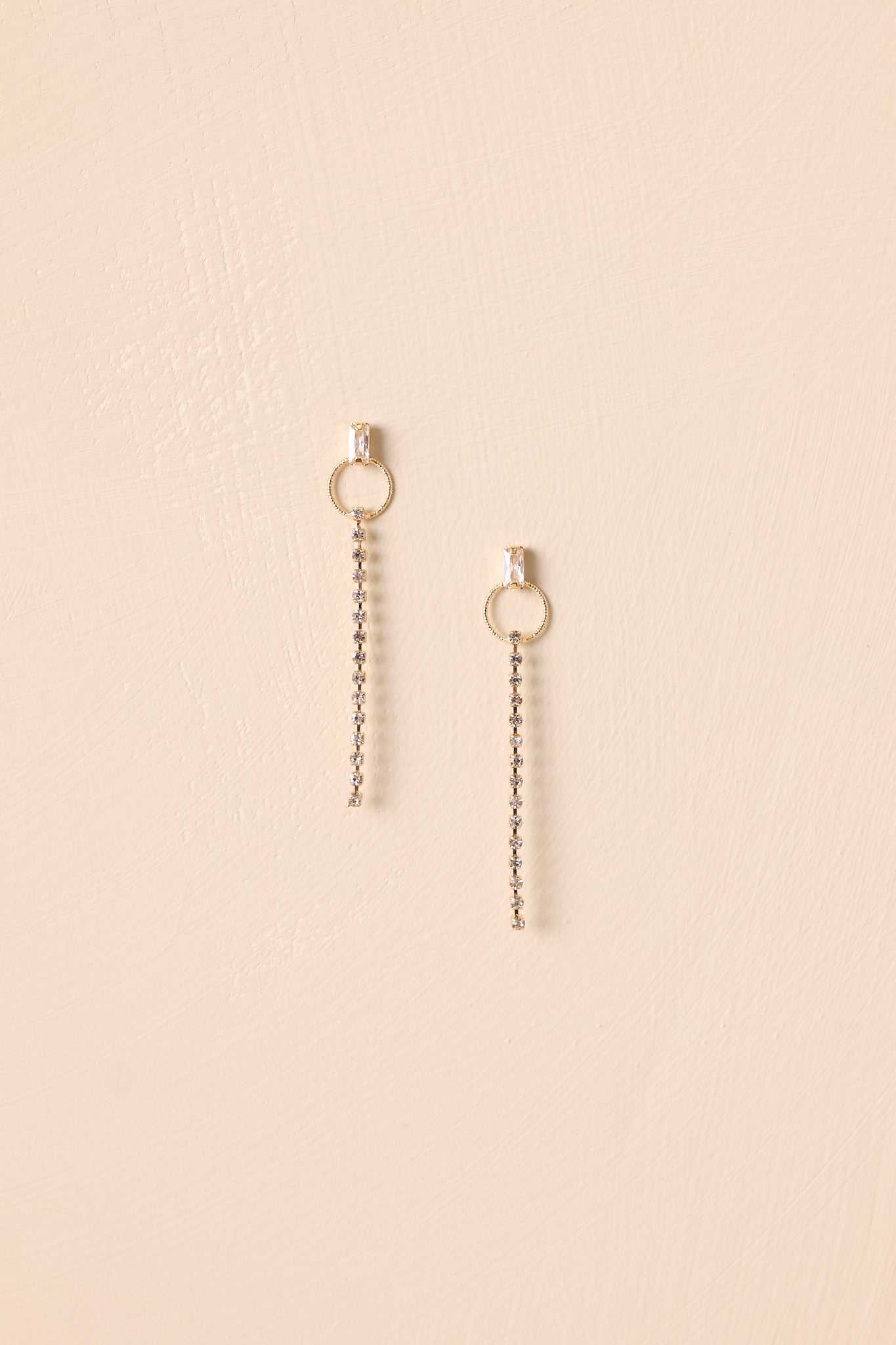 Pair of gold earrings with open circle connector and rhinestone chain drop shown on beige background.