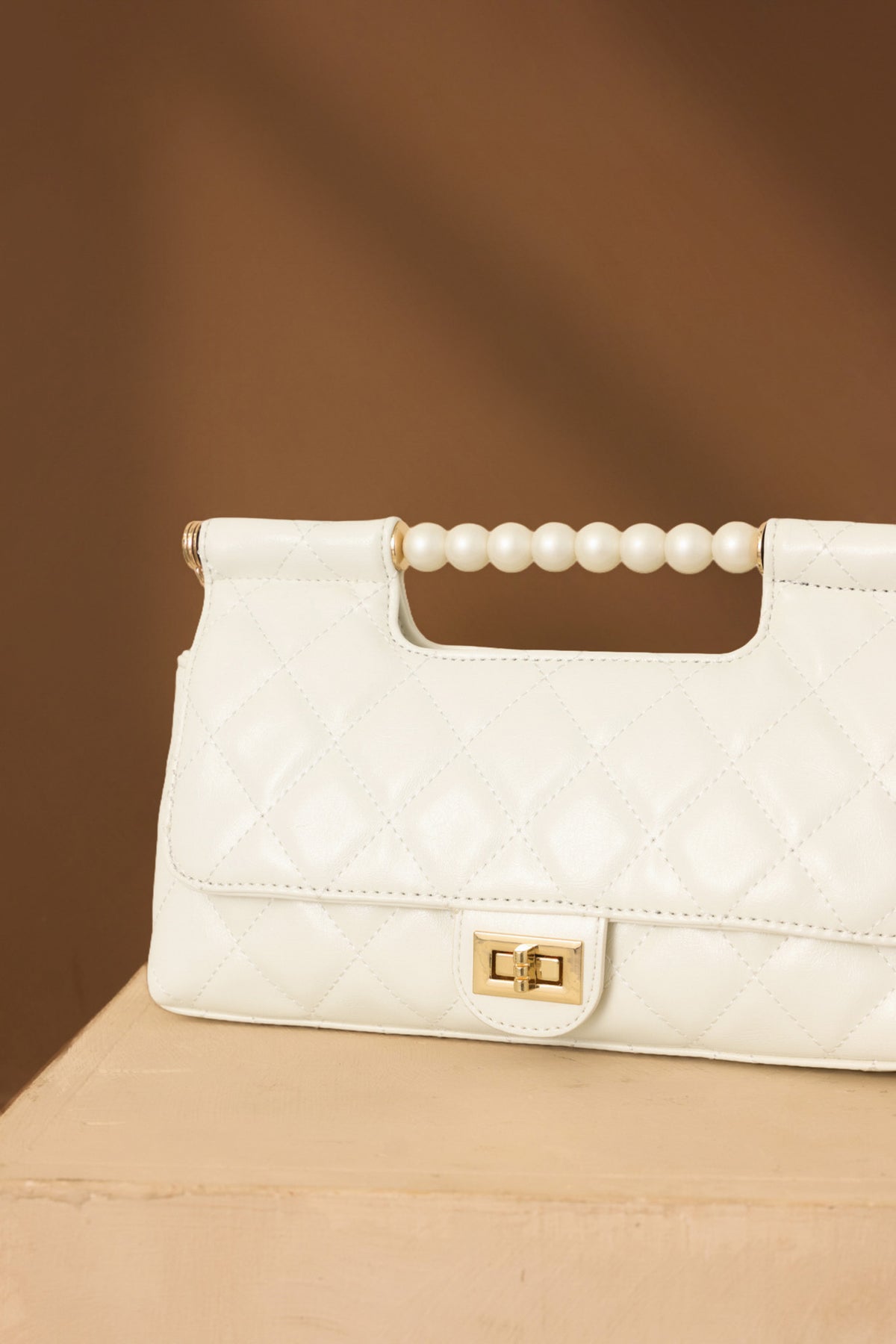Close-up of the handbag revealing the pearl top handle, gold-tone turn lock closure, and smooth faux leather texture on brown back drop.
