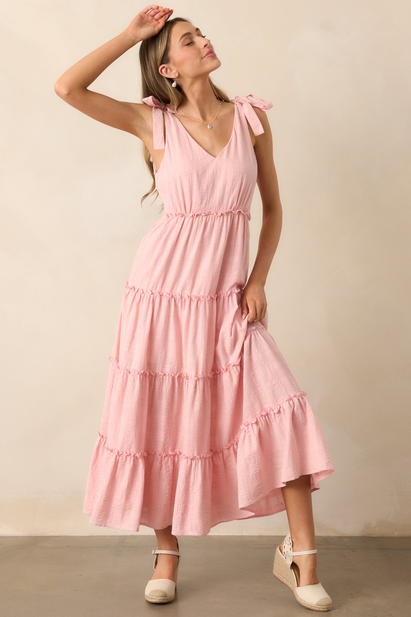 A pink dress with a v-neckline, lightweight textured fabric, and self-tie bow straps, featuring a tiered skirt and ruffle detailing for a feminine touch.