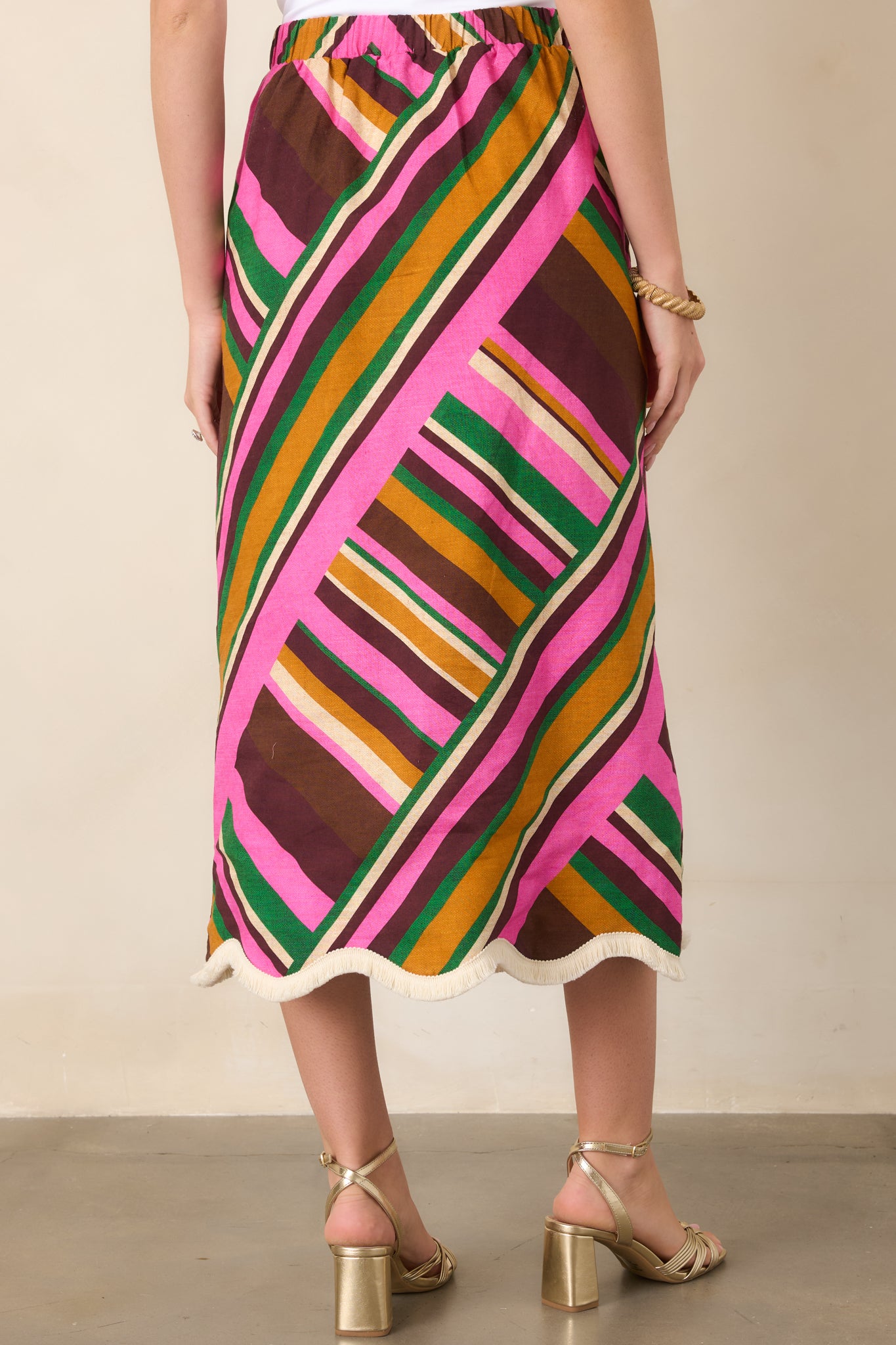 Rear view of the pink geometric striped midi skirt, showcasing its smooth high-rise waistband, softly flared shape, and scalloped fringe-trim hem.
