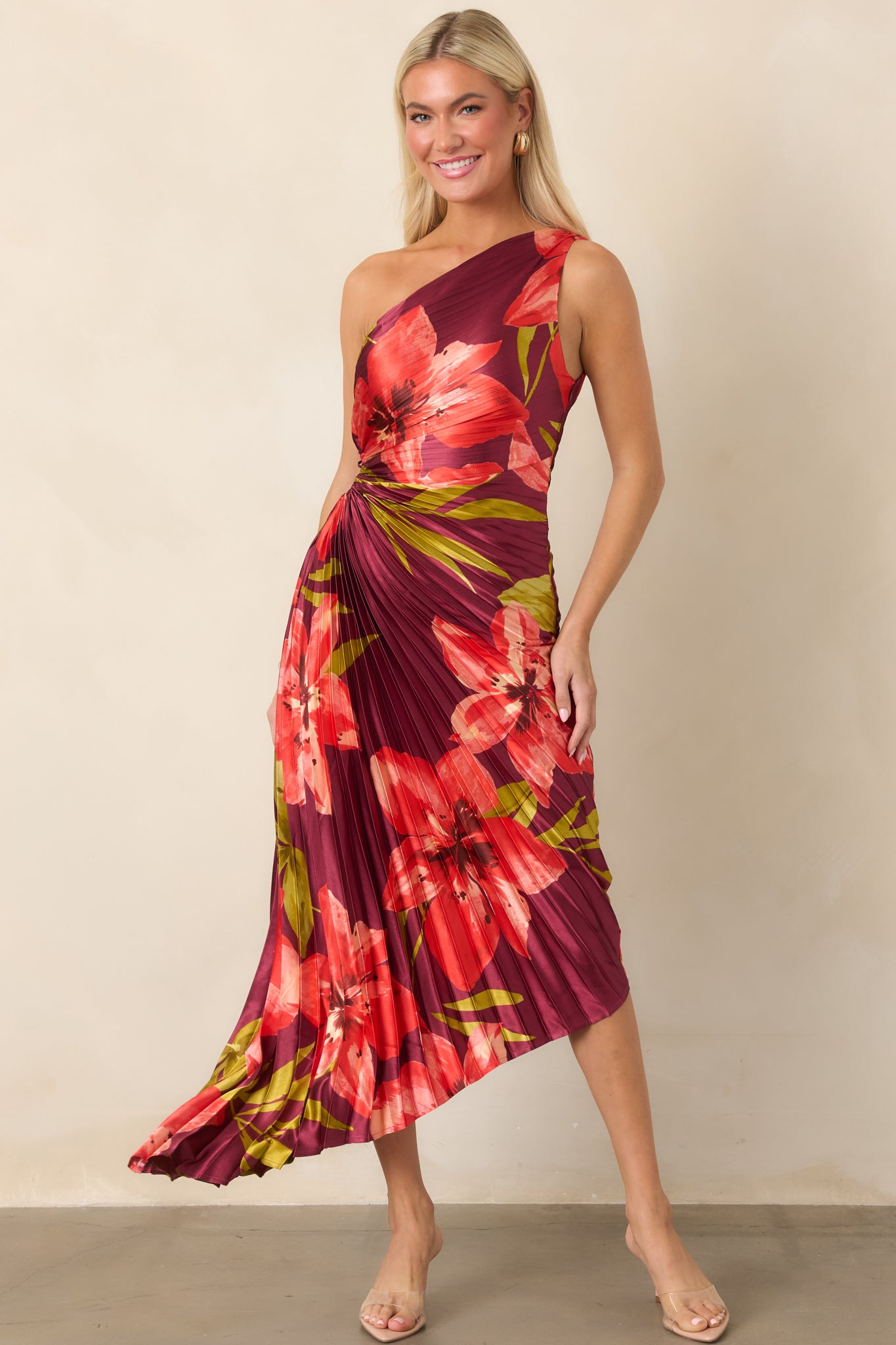 Bold floral satin dress featuring coral and olive hibiscus blooms, dimensional pleats, waist cutout, and flowing uneven hem.