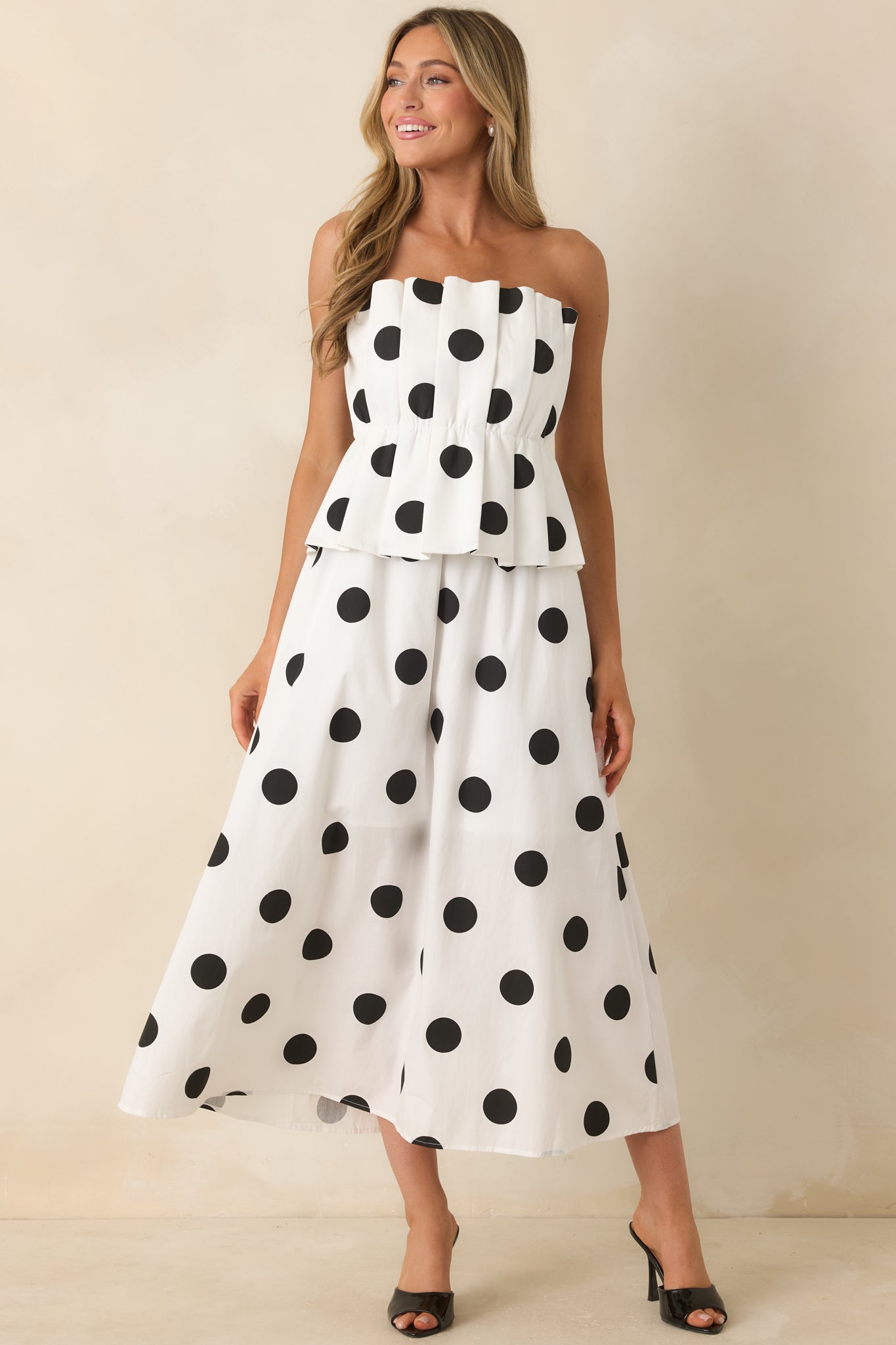 Polka dot midi dress showcasing a strapless bodice, structured pleated overlay, concealed zipper closure, and a softly flared skirt.