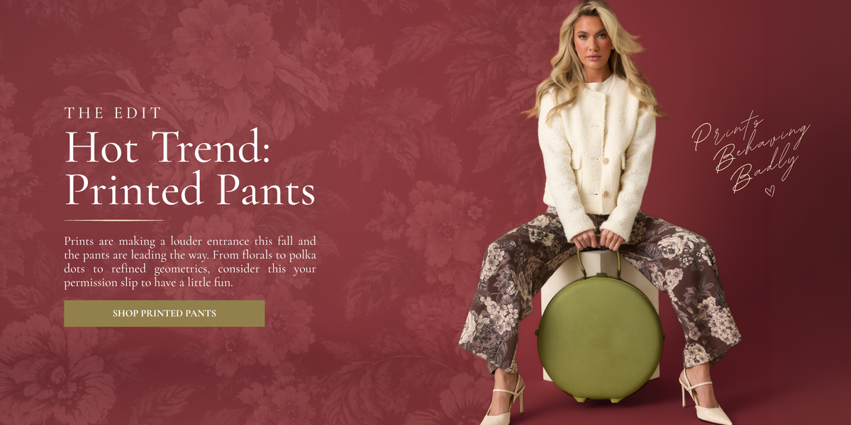 Blog banner featuring a women modeling an ivory jacket, floral printed pants, and sitting down with a green suitcase in front of her. The Blog copy reads, "Hot trend: Printed Pants." It also includes a small description about how printed pants are trending this fall, with a "Shop Printed Pants" button. 