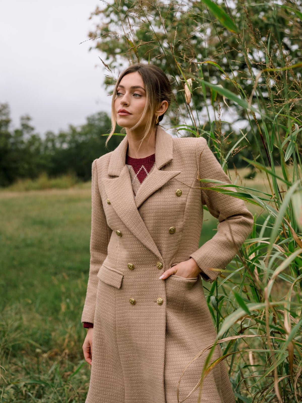 Woman in a long camel double-breasted tailored topcoat with gold buttons standing beside tall grass.