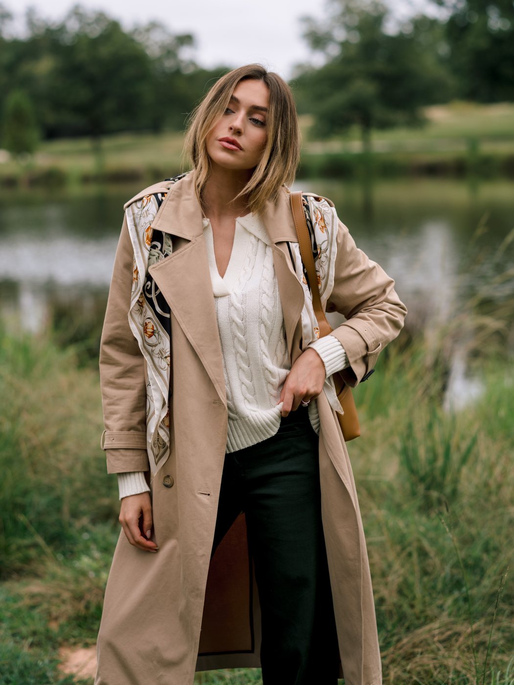 Woman wearing an open tan trench coat over an ivory cable knit and dark trousers in a grassy field.