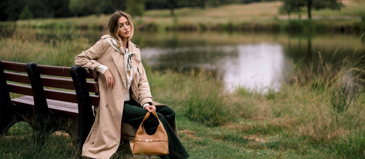 Woman in a tan belted trench coat and ivory knit sitting on a park bench by a pond, holding a tan leather bag.