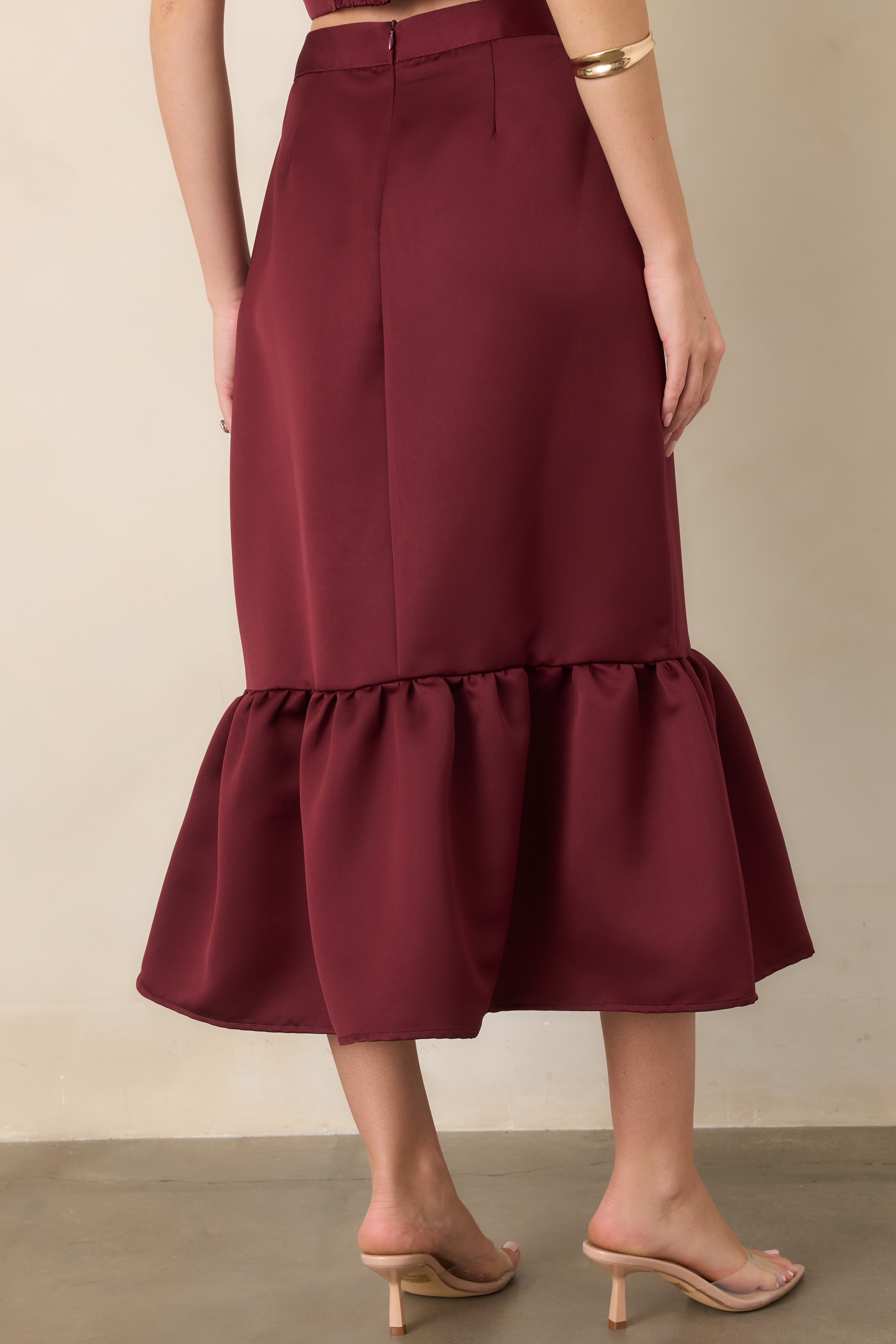 Rear view of the burgundy satin midi skirt showcasing its fitted silhouette, smooth finish, and gathered flounce hem.
