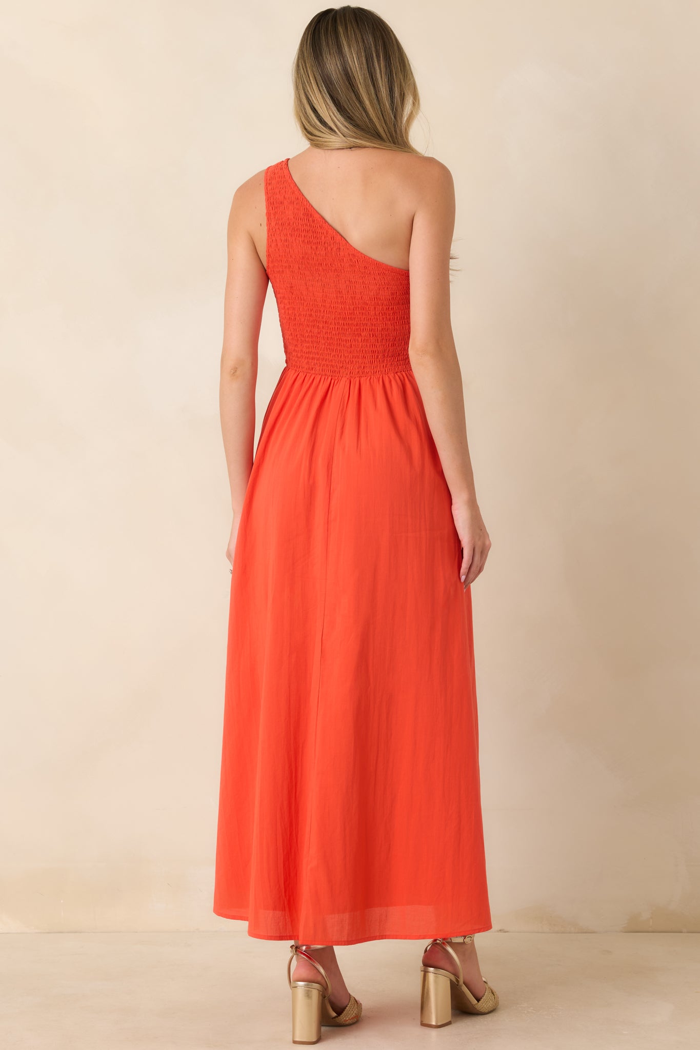 Rear view of the coral-orange cotton maxi dress showcasing the fully smocked bodice, one-shoulder silhouette, and flowing A-line skirt.