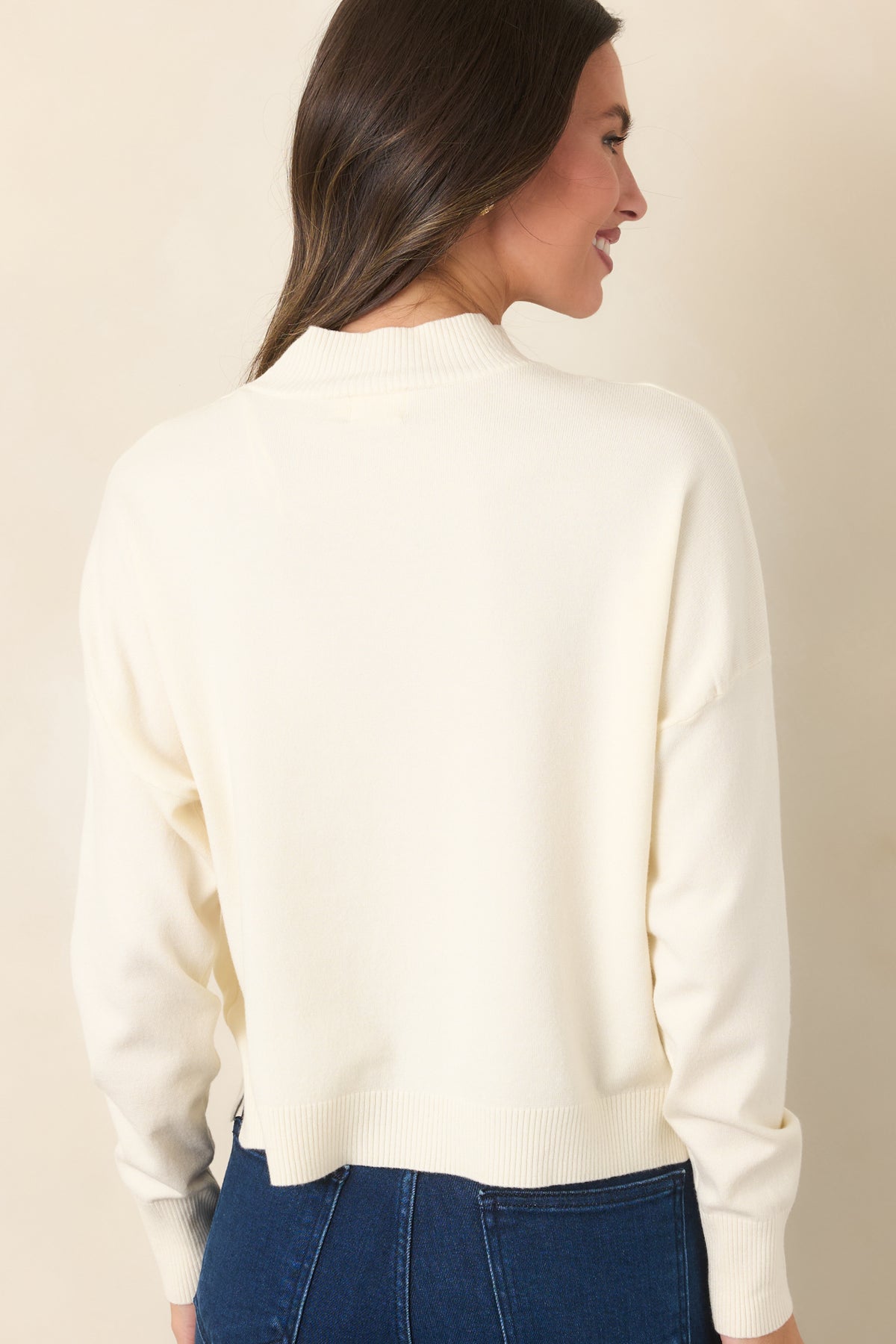 Rear of the ivory knit pullover featuring a simple back panel, dropped shoulders, long sleeves, and a ribbed hem with side slits.