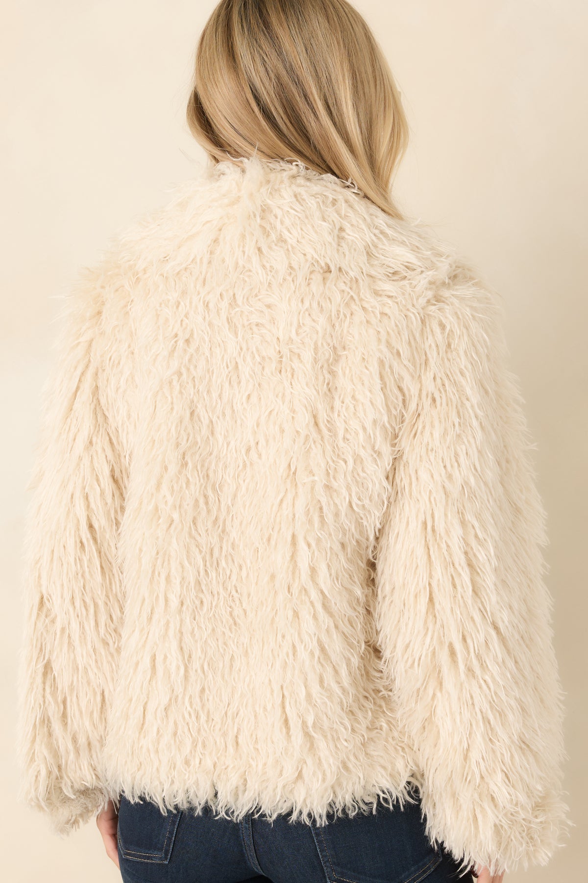 Rear view of the neutral plush jacket showing its shaggy wavy texture, long sleeves, and relaxed hip-length silhouette.
