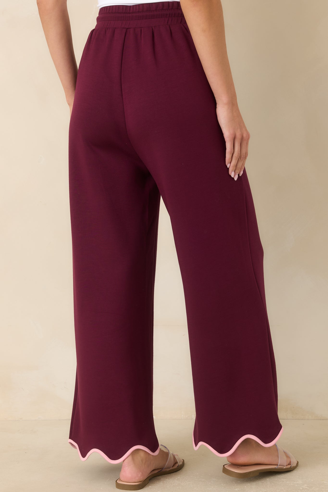 Rear view of the burgundy knit pants showcasing the flowing wide leg silhouette and blush pink scalloped trim along the hem.