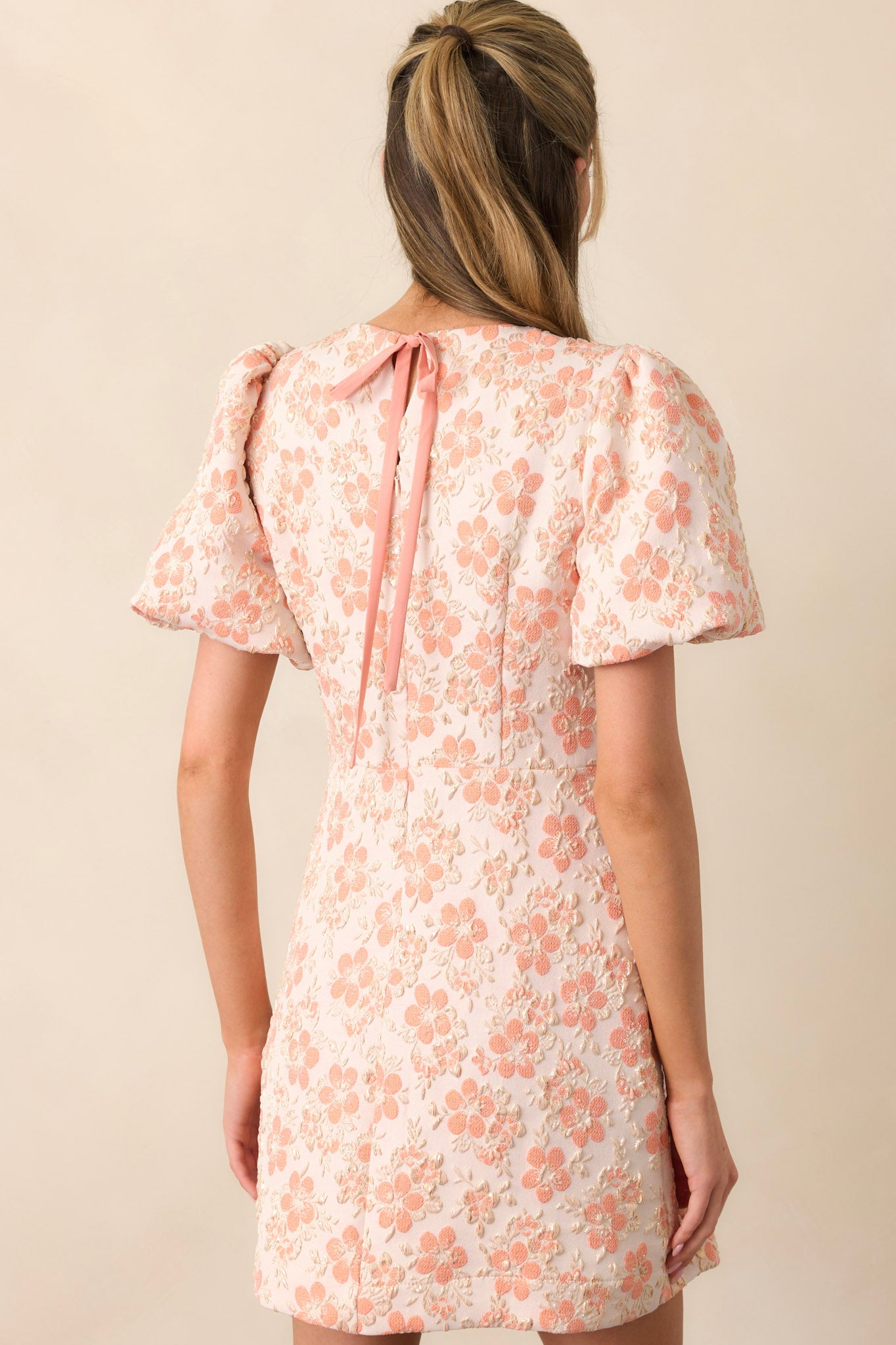 The rear view of an apricot mini dress, featuring a discrete zipper and a keyhole self-tie closure at the neckline. The puff sleeves and gold detailing continue seamlessly throughout the design.