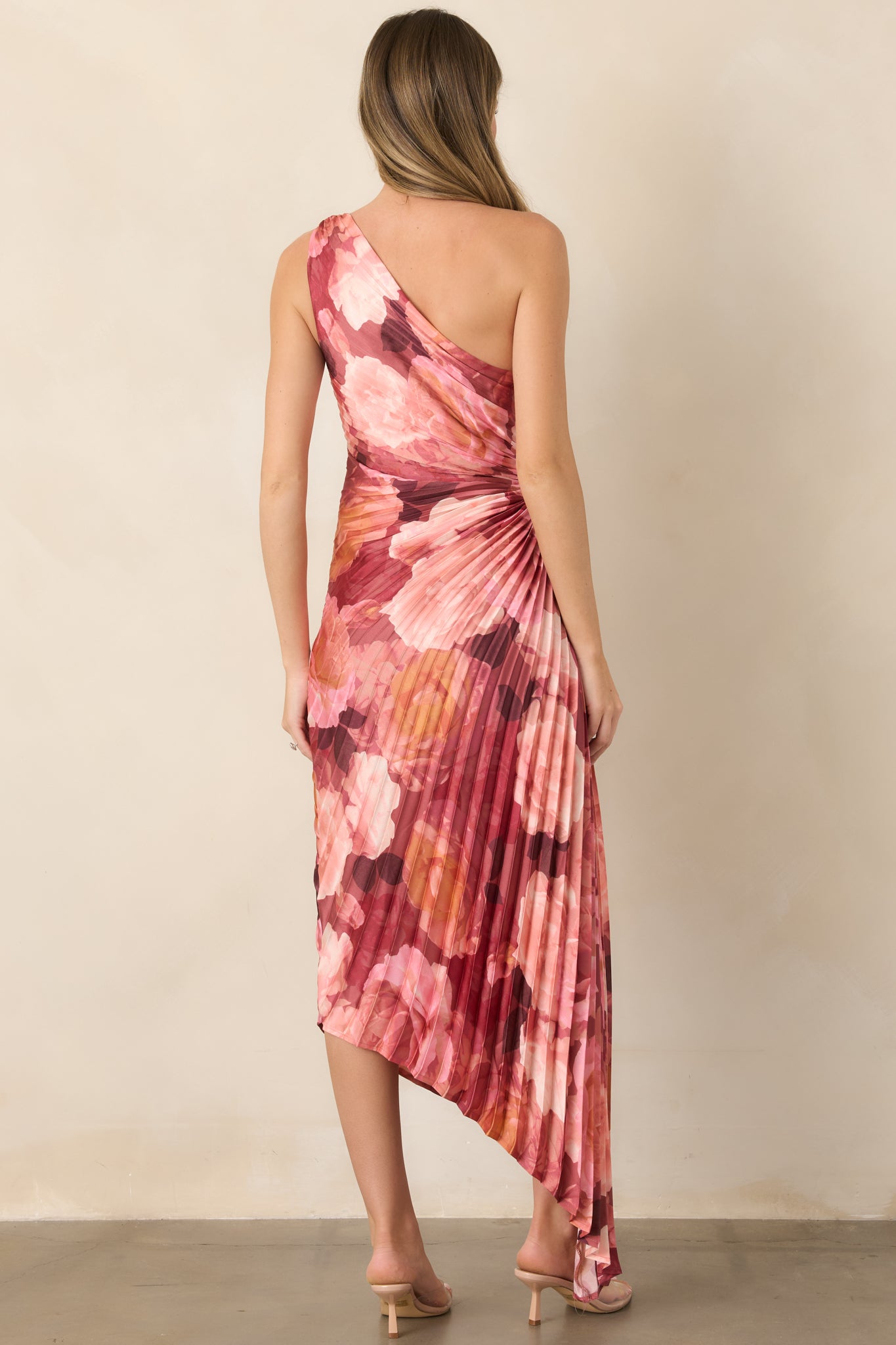 Rear view of the floral satin maxi dress displaying its pleated fabric, fitted waist, flowing asymmetrical skirt, and pink, rose, and burgundy floral pattern.