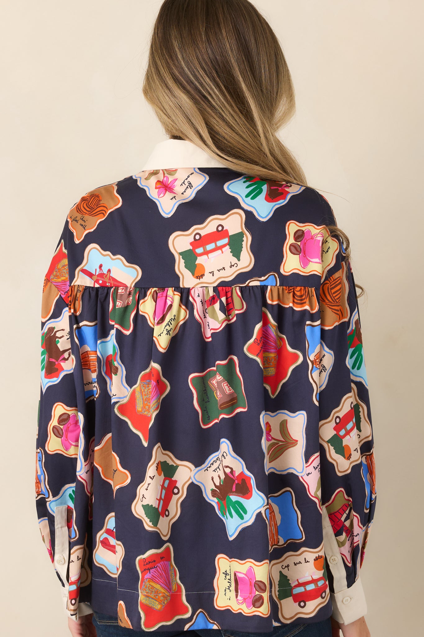 Rear view of the midnight blue printed top featuring its gathered back yoke, ivory cuffs, and colorful illustrated stamp motifs.