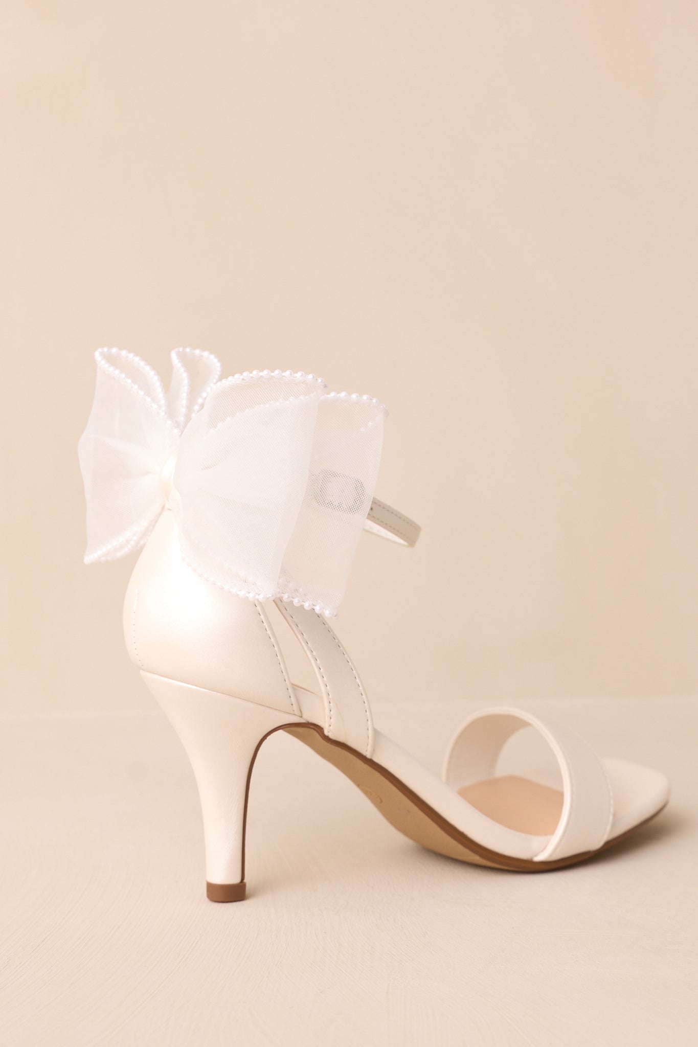 Rear view of the white heels, highlighting the lace bow with pearls and the thin heel, along with the ankle strap seen from behind.