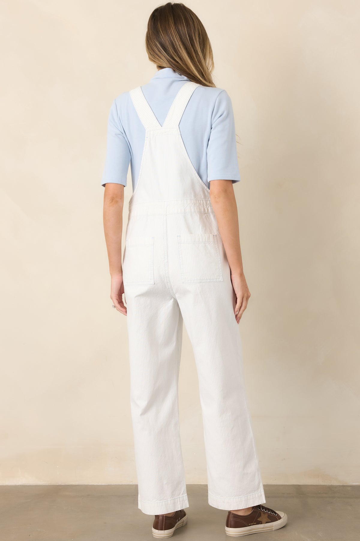 Rear view of the ivory and sky blue striped cotton overalls showcasing the adjustable cross-back straps, side buttons, straight-leg silhouette, and ankle-length hem.