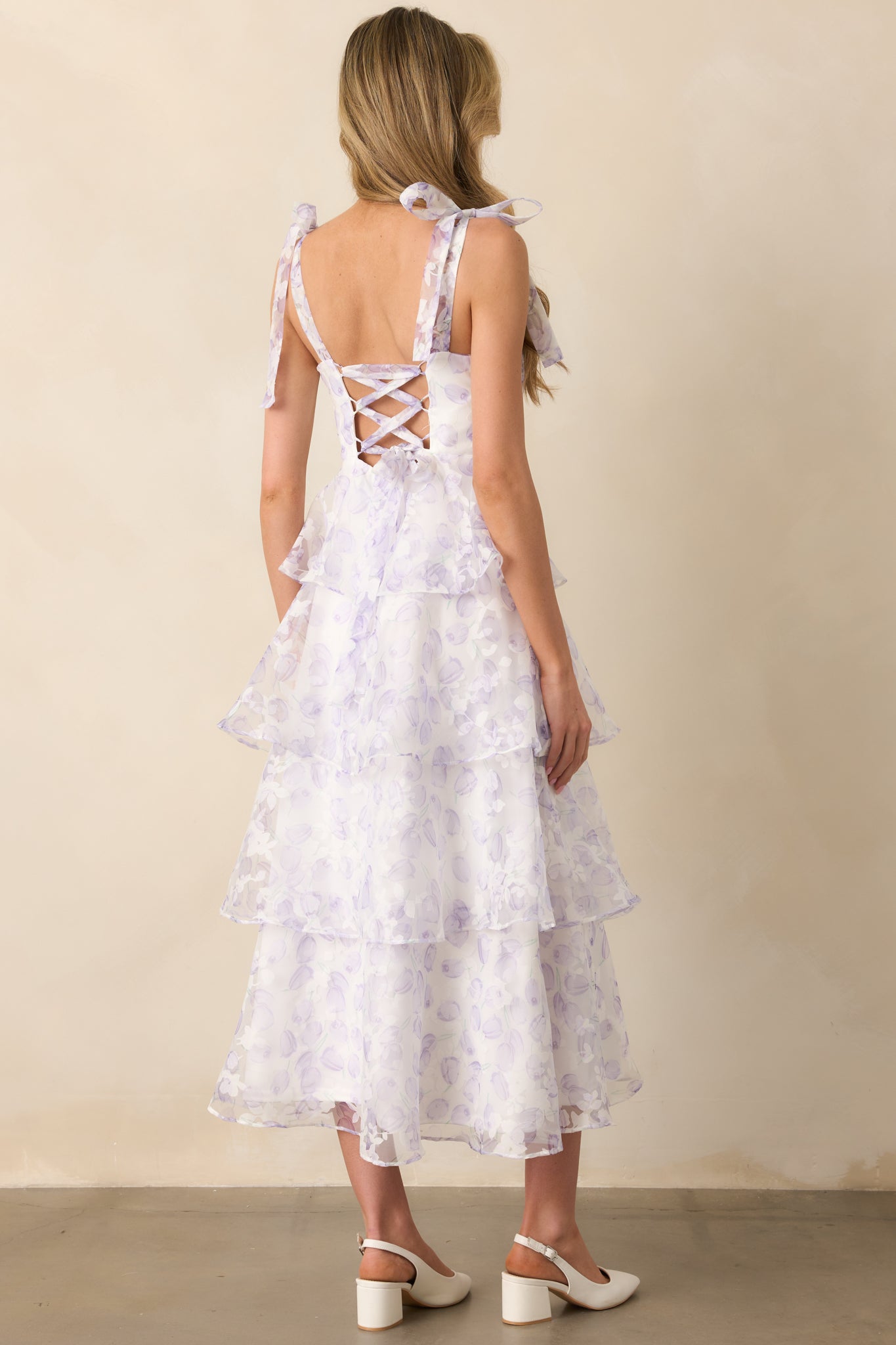  The rear view of a lavender midi dress featuring an adjustable self-tie lace-up corset, a hidden zipper closure, and a floral tulle overlay. The tiered skirt cascades gracefully for a soft, elegant finish.