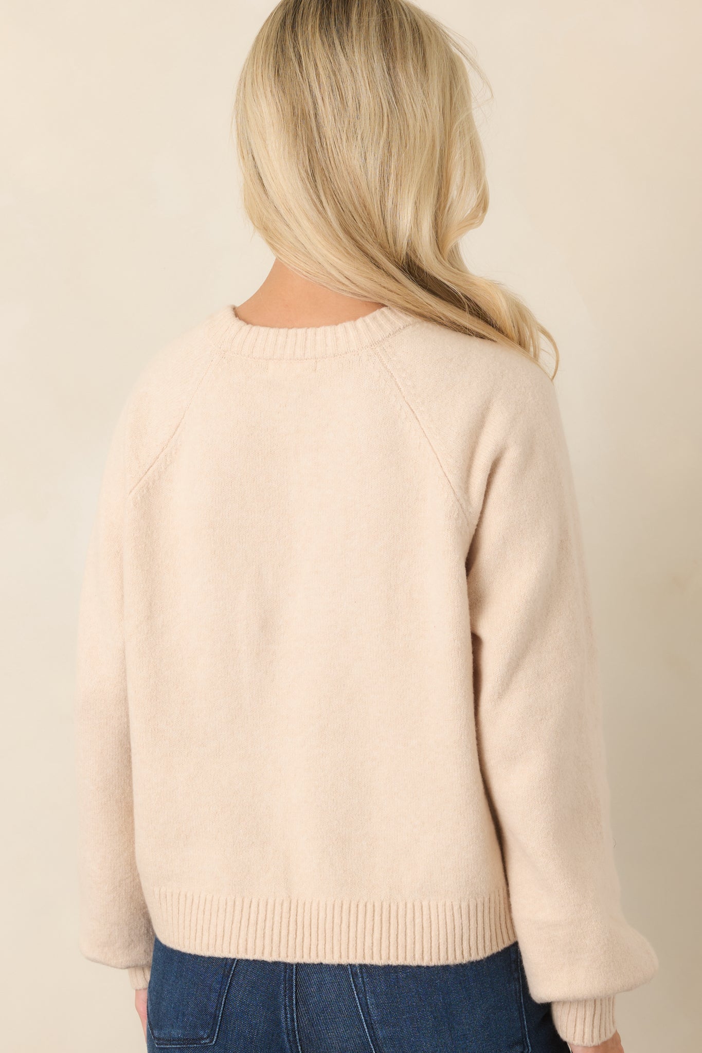 Rear view of the soft ecru knit top showcasing the slightly cropped silhouette, long raglan sleeves, gathered shape, and ribbed hem.