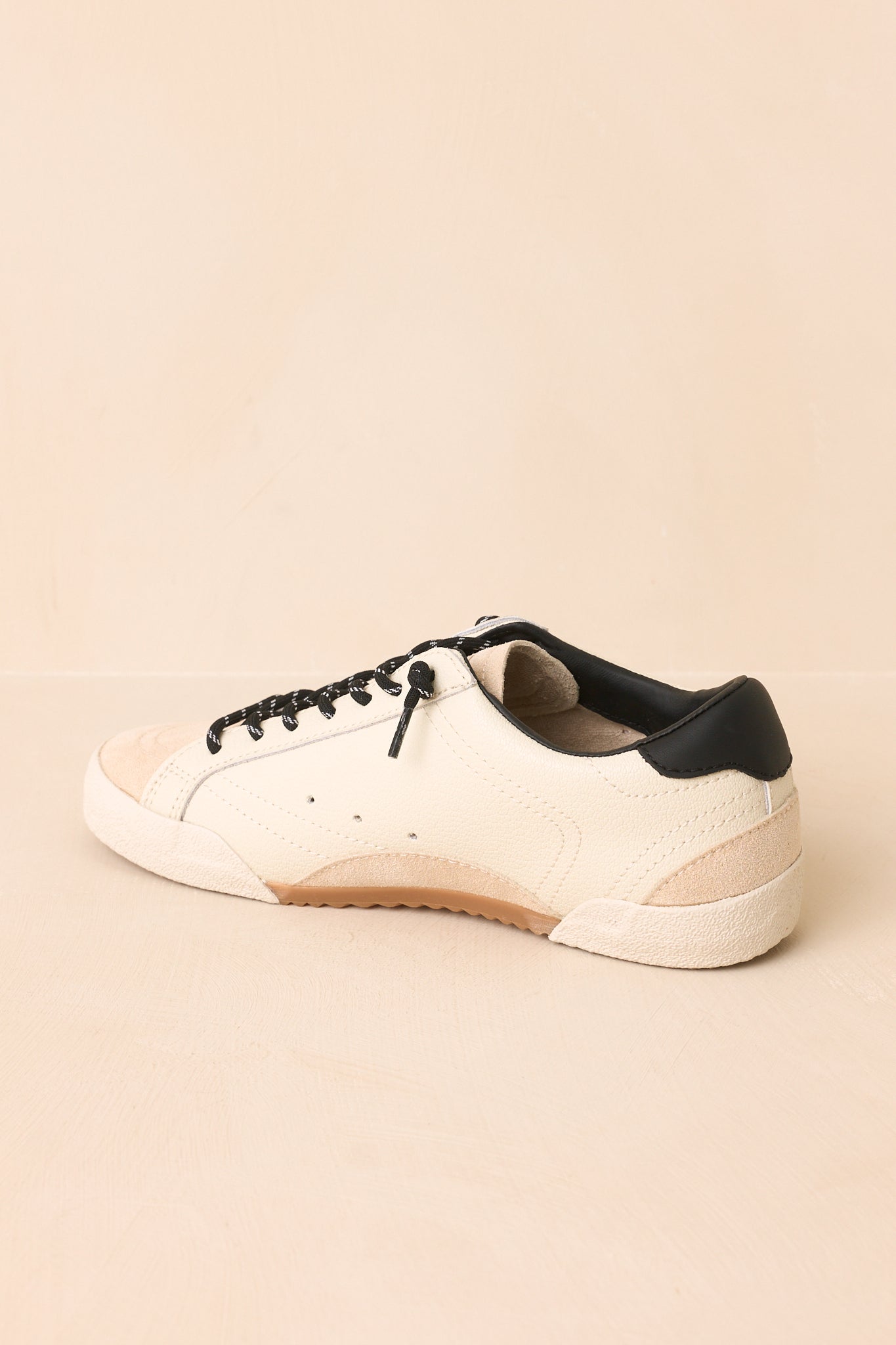 Rear view of the white sneakers highlighting the black heel accents, low-profile shape, textured soles, and vintage-inspired finish against a beige background.