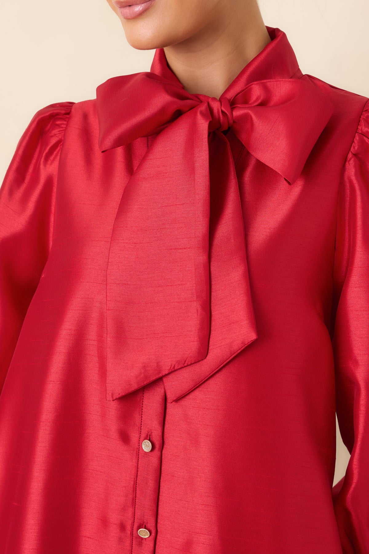 Close-up of the red dress showing the oversized bow neckline, lustrous fabric, and gold-tone front buttons.