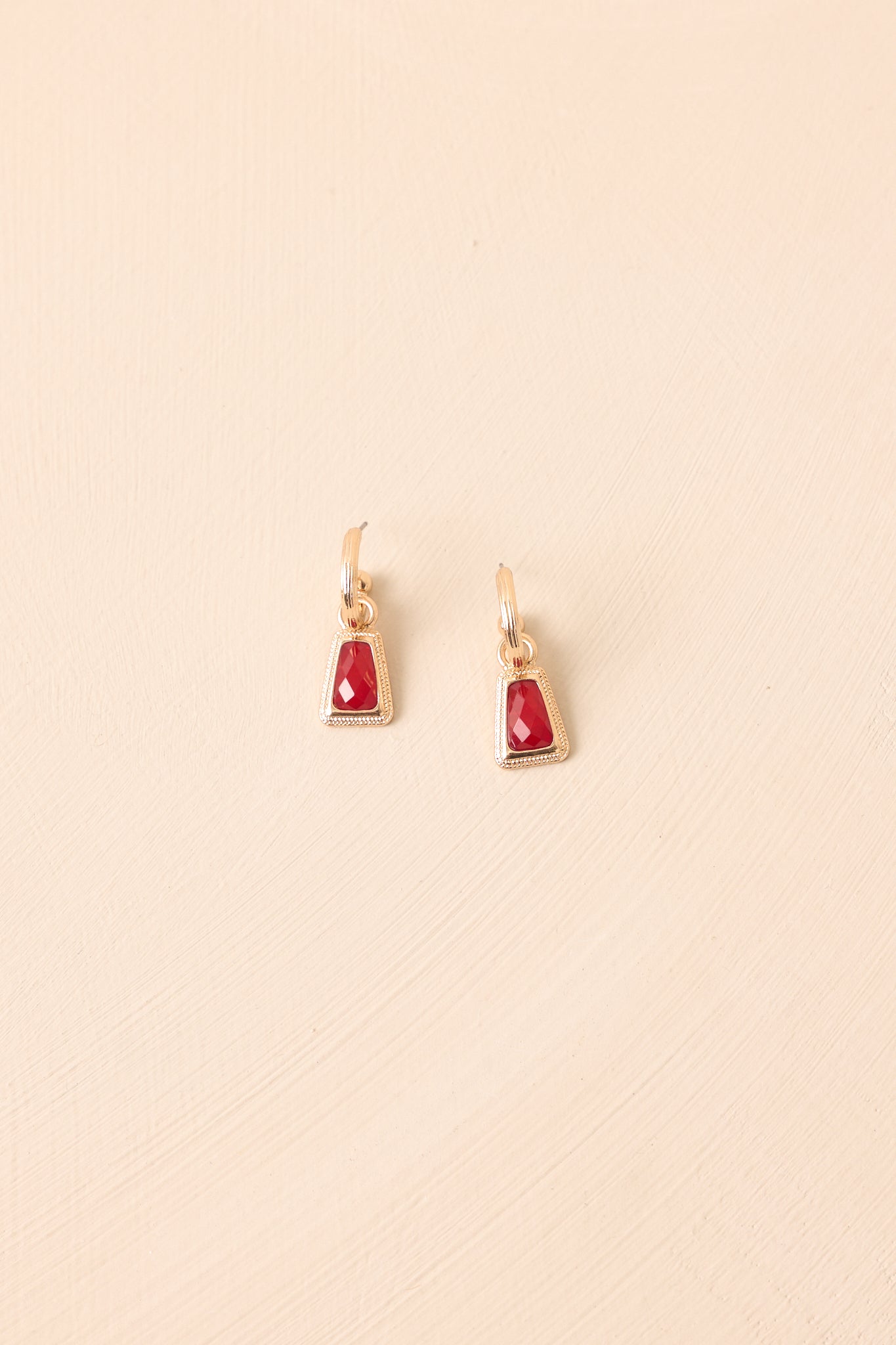 Red stone drop earrings displayed against a beige background, emphasizing the trapezoid gold texture and clasp design.