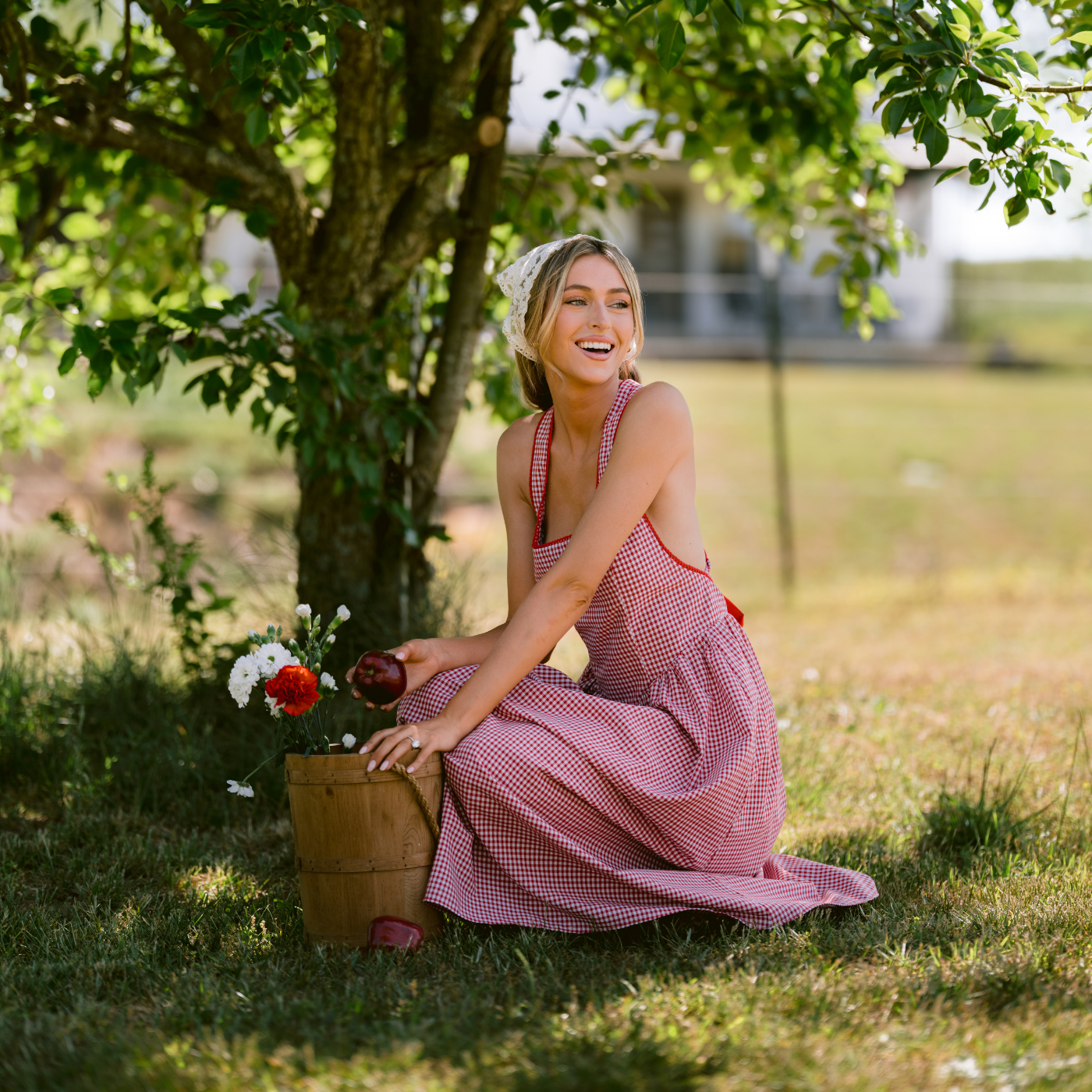Woman in a red, gingham dress sitting under a tree with flowers and a bucket of apples.