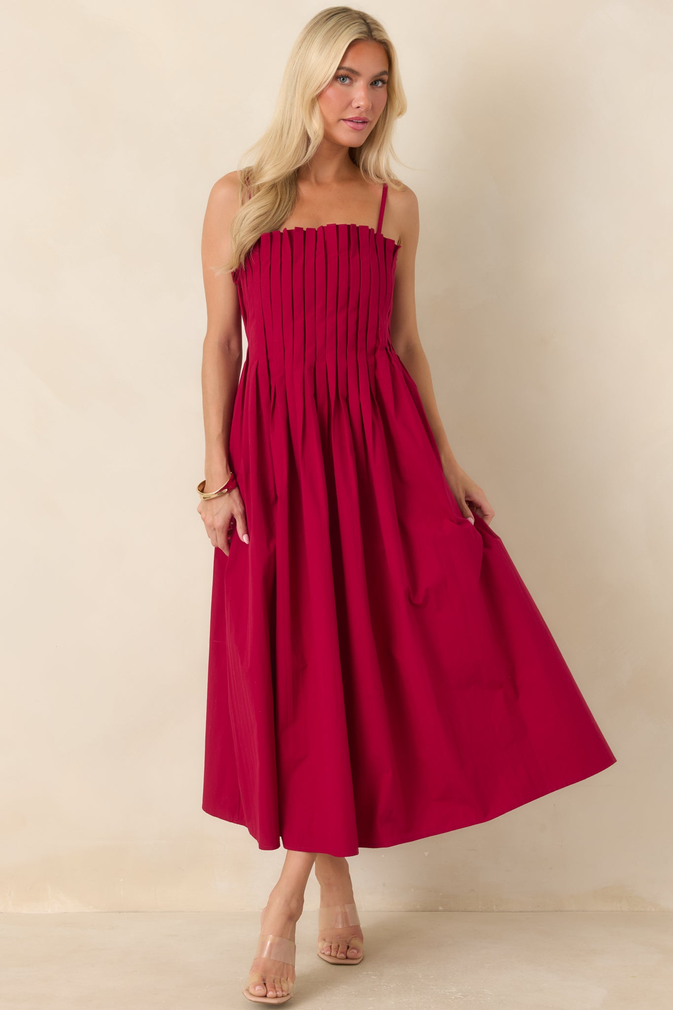 Ruby red midi dress designed with a square neckline, crisp vertical pleats through the bodice, adjustable straps, and a full skirt that creates graceful movement.