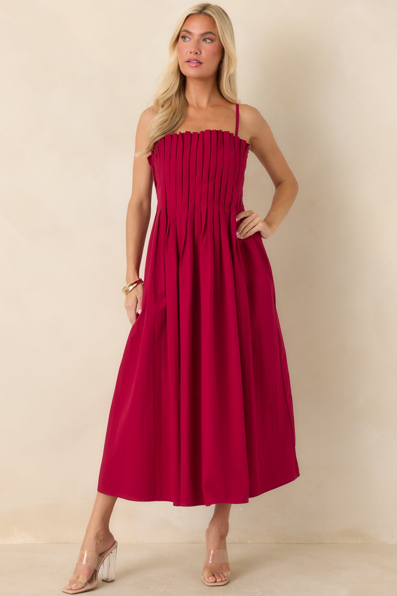 Elegant ruby red cotton dress featuring a fitted pleated bodice, adjustable spaghetti straps, practical side pockets, and a softly gathered midi-length skirt.