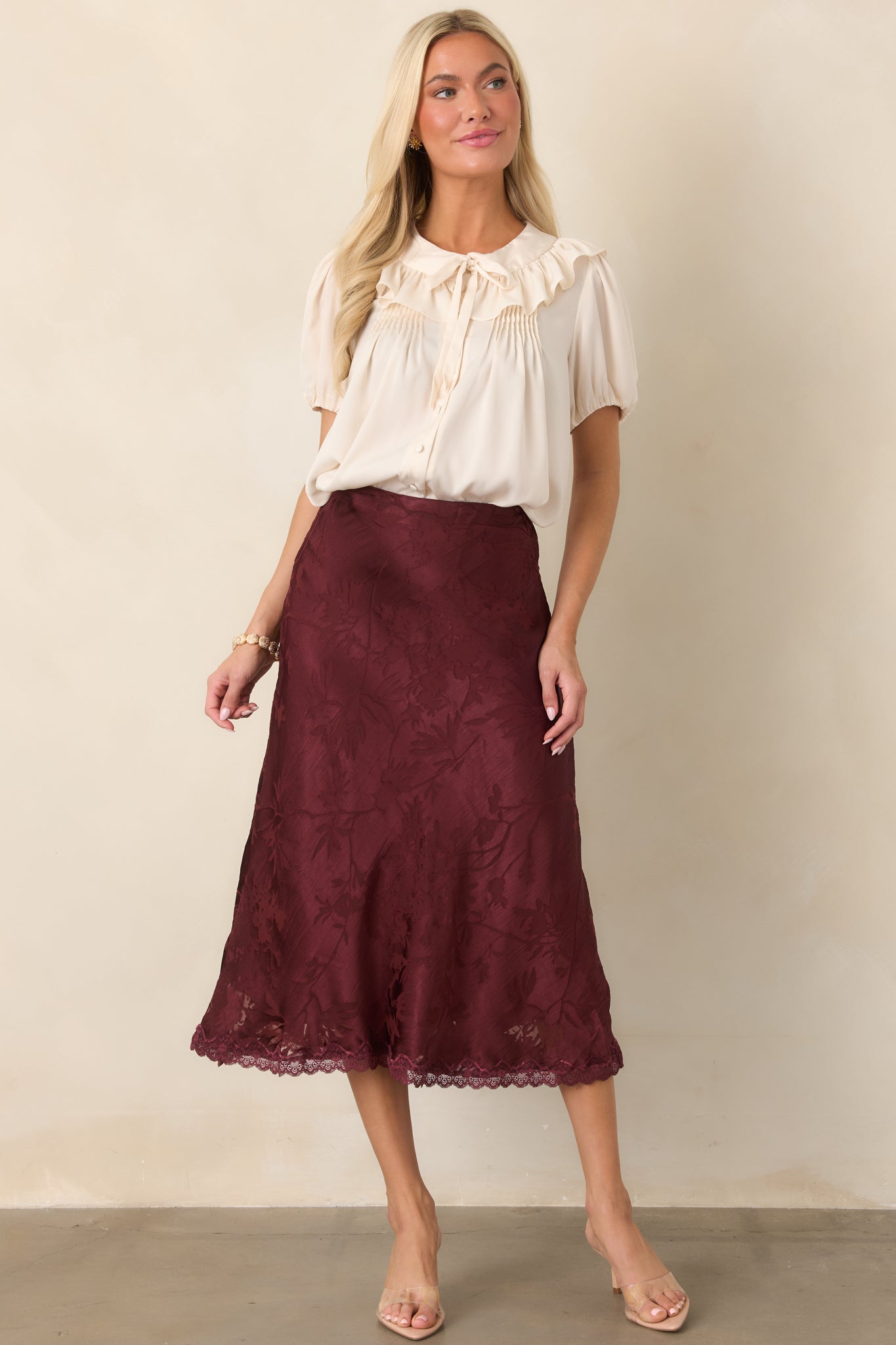 Feminine cropped blouse highlighting the wide ruffled collar, tied neckline, covered buttons, and gathered construction in a warm champagne hue.