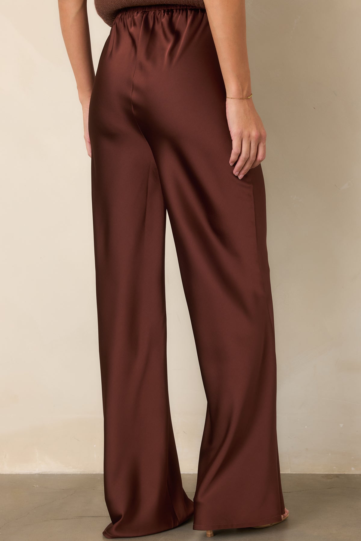 Rear of the rust brown satin pants showcasing the elasticized waistband, fluid full-length legs, and flowing wide-leg silhouette.