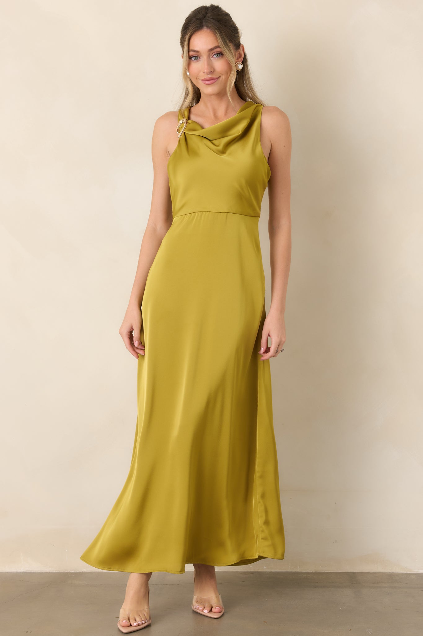 Satin maxi dress with a softly draped neckline, sculptural gold-tone shoulder accent, empire waist, and flowing A-line silhouette.