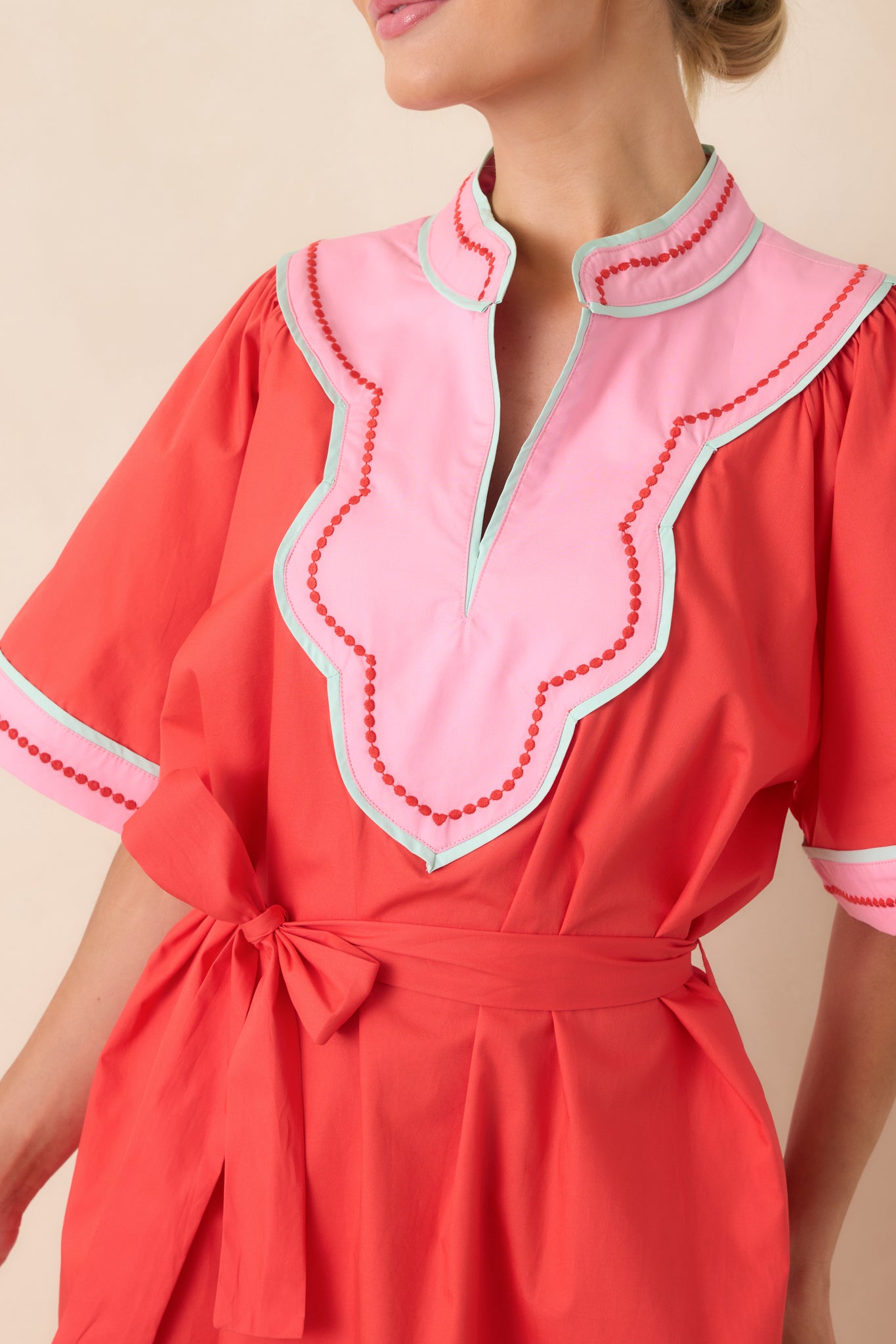 Detailed view of the scalloped V-neckline and pink contrast bib panel, highlighting the crisp cotton fabric and bold trim against the poppy red base.