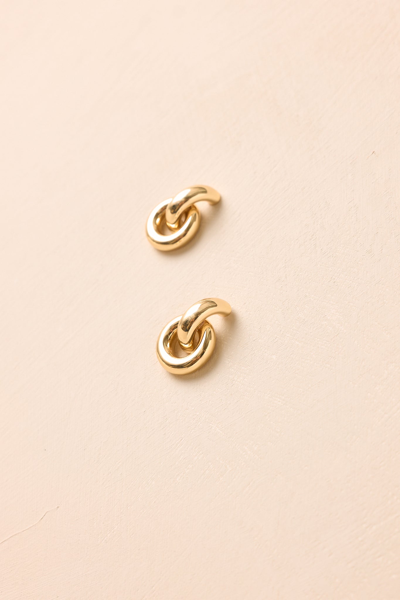 Sculptural gold earrings presented on a neutral beige backdrop, showcasing layered circular shapes and a reflective high-shine surface.