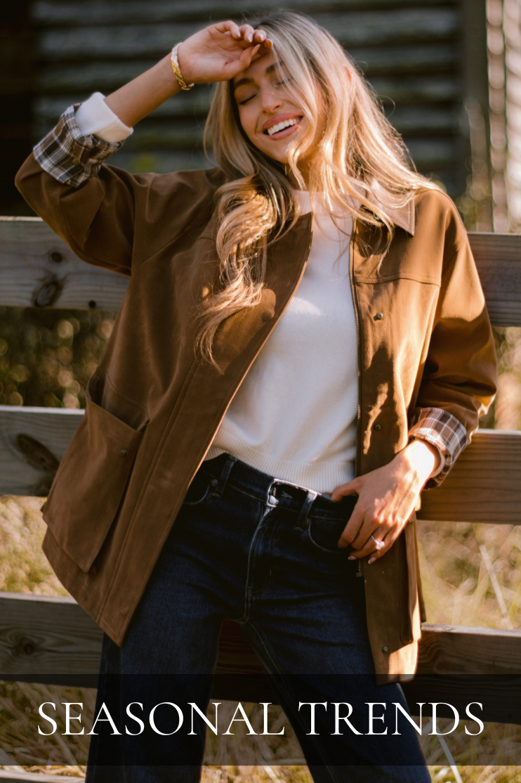 Model smiling outdoors in a casual fall outfit featuring a brown jacket with plaid cuffs, white sweater, and dark denim jeans. Captures effortless autumn style from the Red Dress Seasonal Trends collection — cozy layers and timeless pieces perfect for fall fashion.