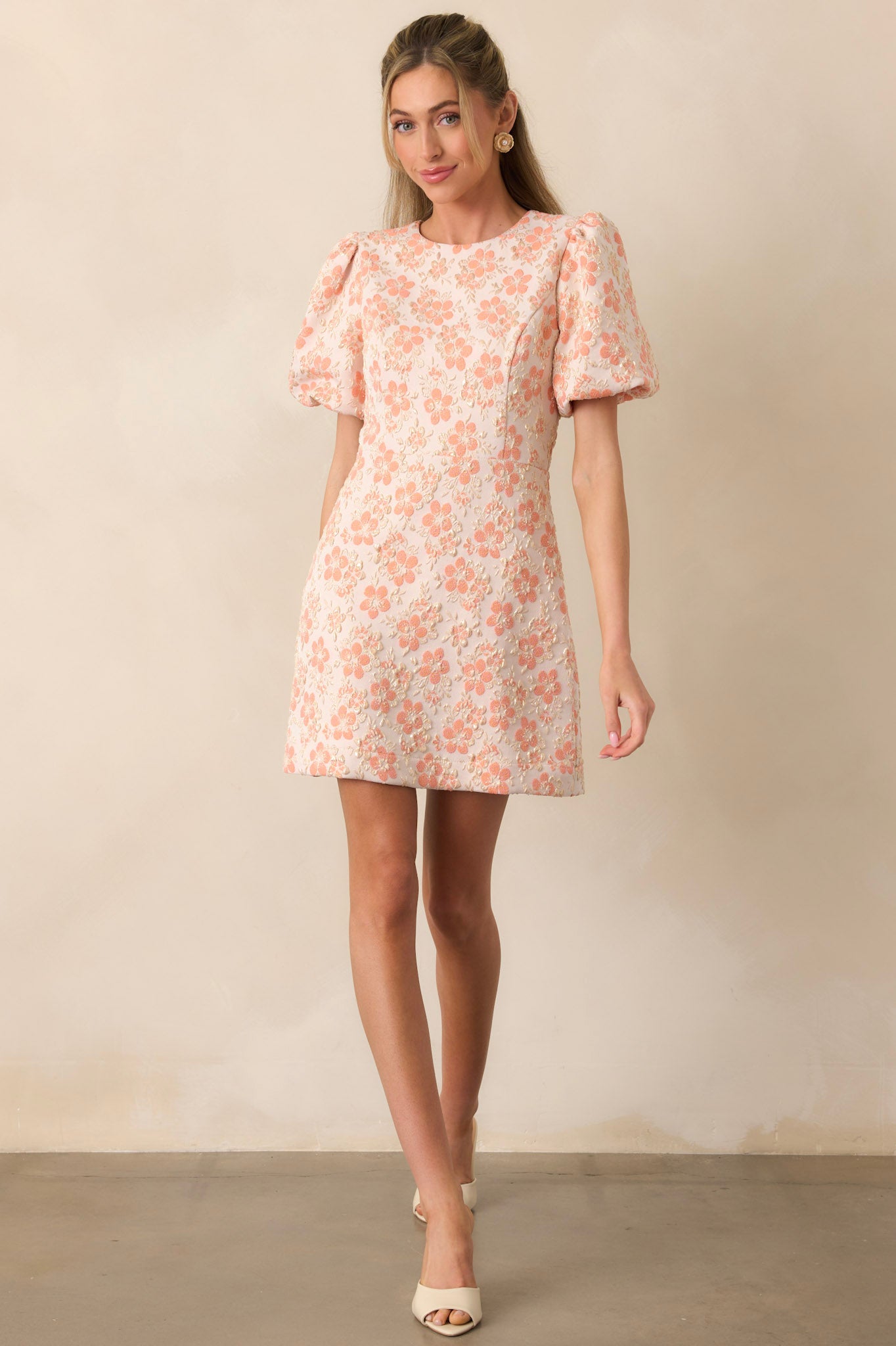A short dress featuring soft apricot fabric, delicate gold accents, and voluminous puff sleeves. The round neckline creates a classic and elegant look.