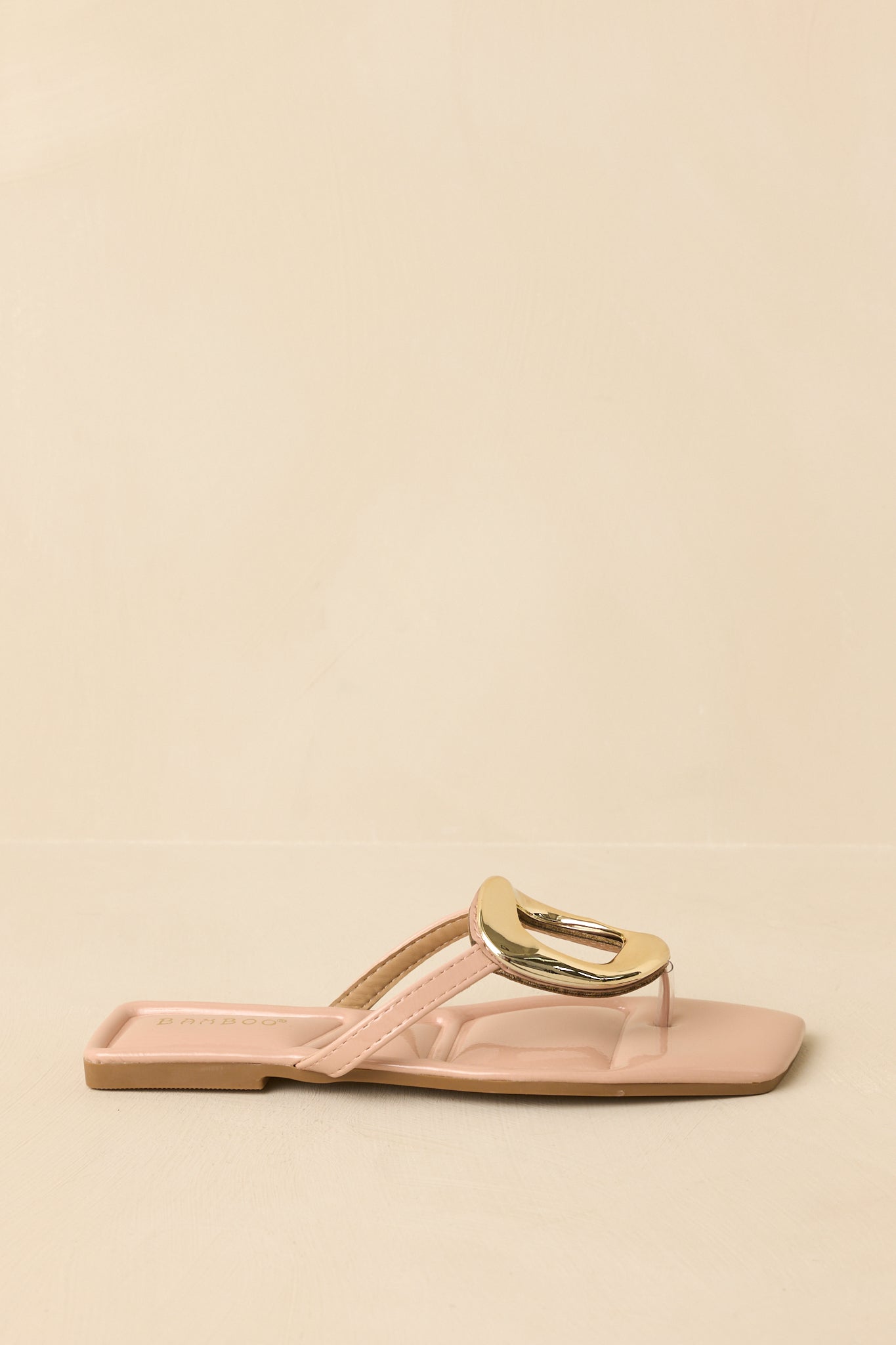Side profile of the nude patent sandals displayed against a beige background emphasizing the flat sole and streamlined shape.