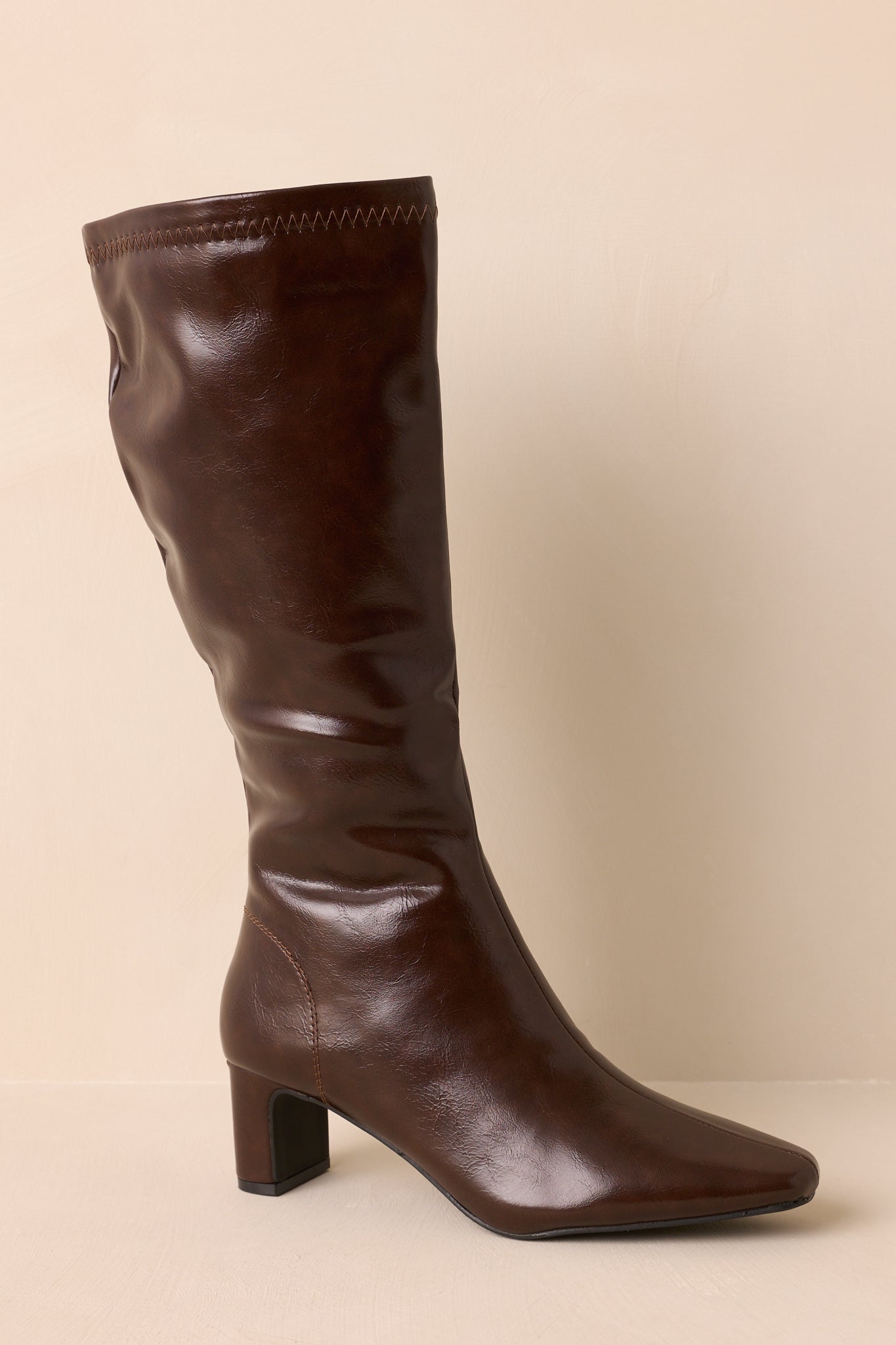 Side view of brown faux leather boots with pointed toe and block heel on a beige backdrop.