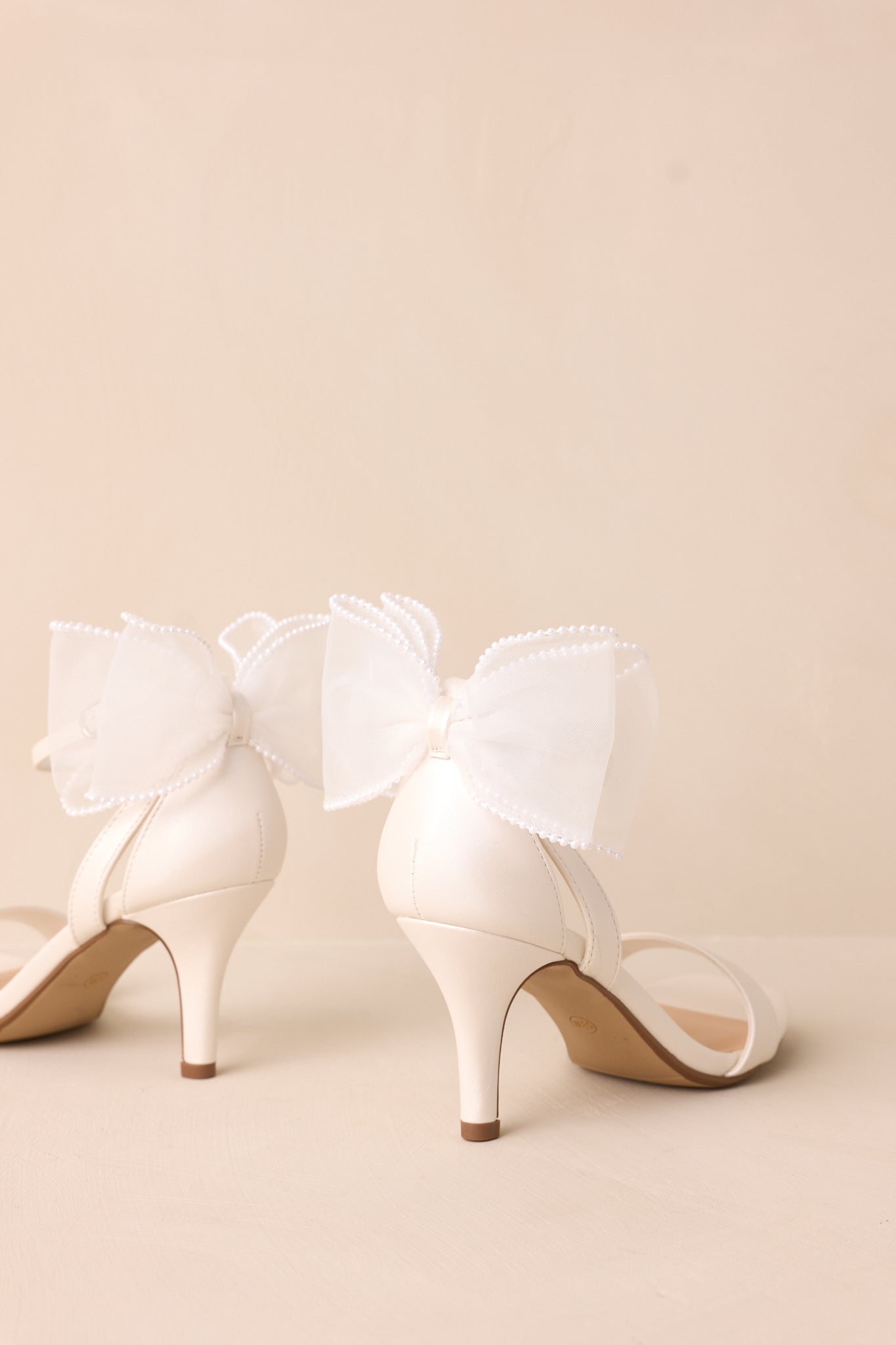 Slightly elevated side view of the white heels, emphasizing the thin heel and the ivory lace bow bordered with small pearls.