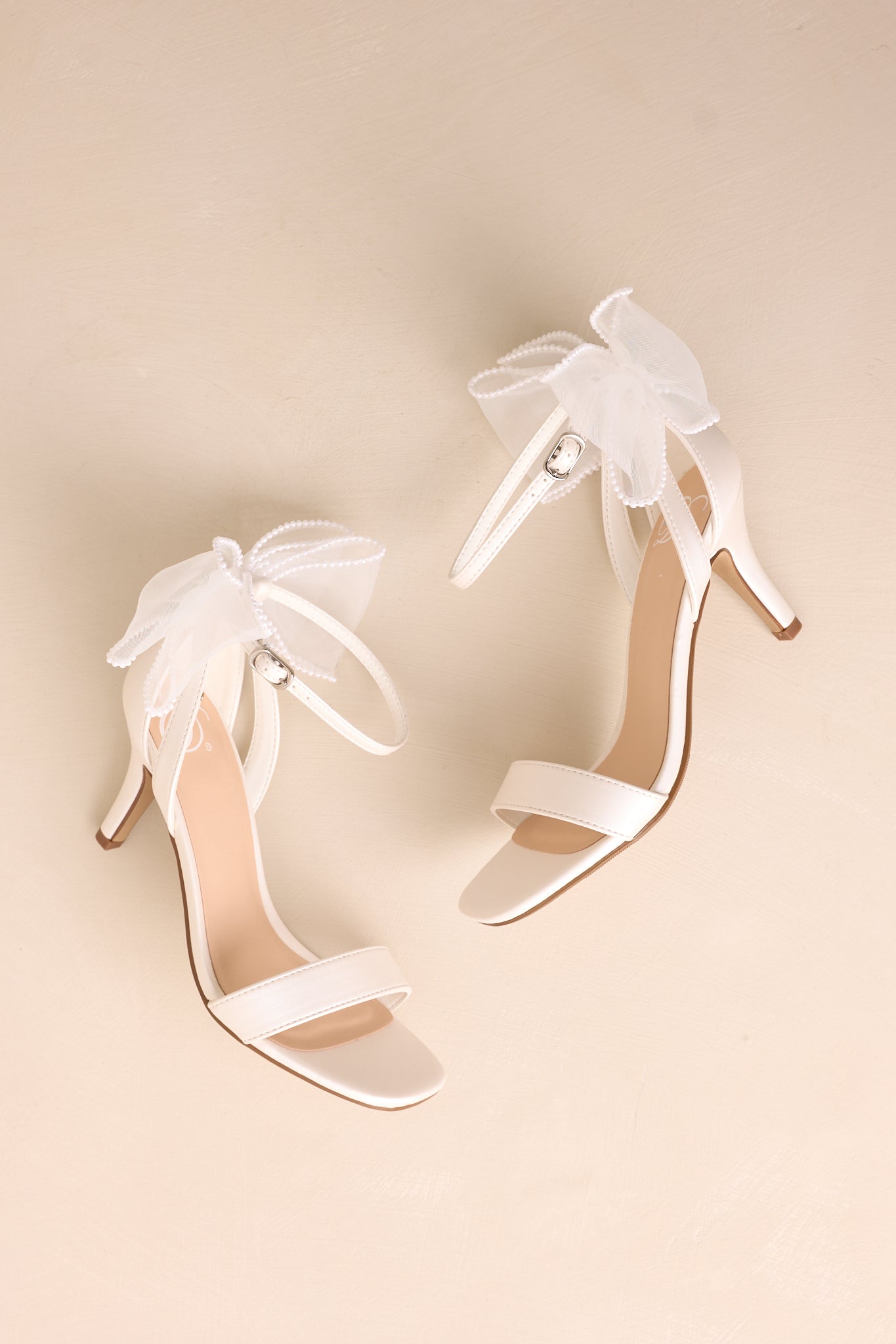 Full side view of the white heels, capturing the smooth white material, ankle strap, and the large ivory lace bow with pearls on the heel.