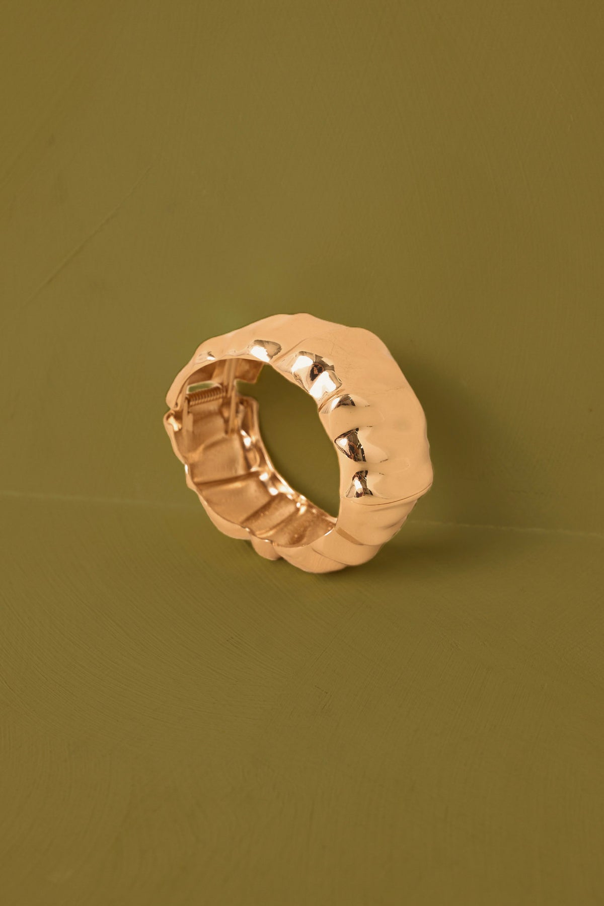 A textured, chunky metallic ring in a warm rose gold finish, photographed on a solid olive green background.