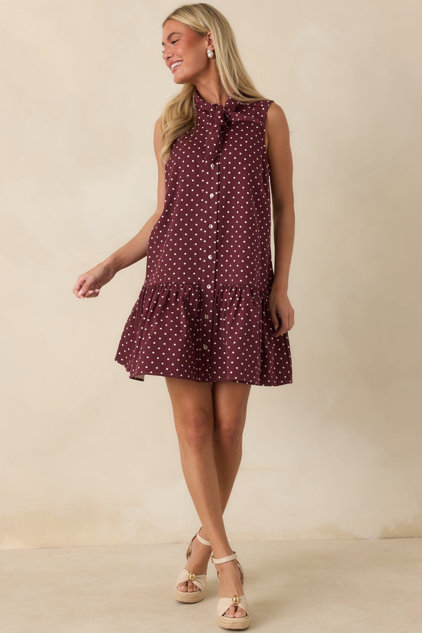 Sleeveless cotton mini dress featuring a self-tie neckline, scattered ivory polka dots, side pockets, and a gathered ruffle hem.