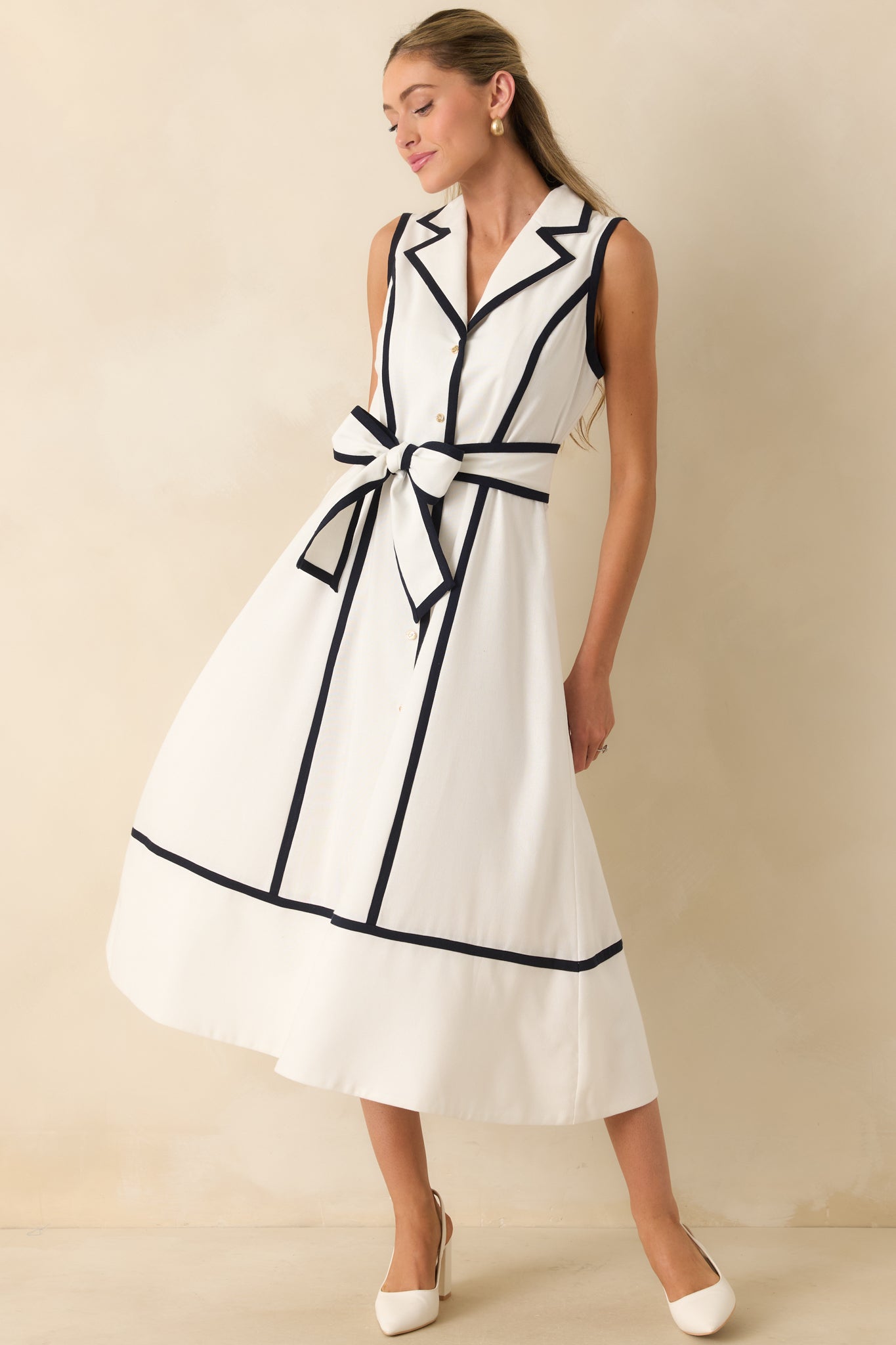 Sleeveless midi dress featuring a notched collar, button-front design, navy contrast piping, and a coordinating self-tie belt.