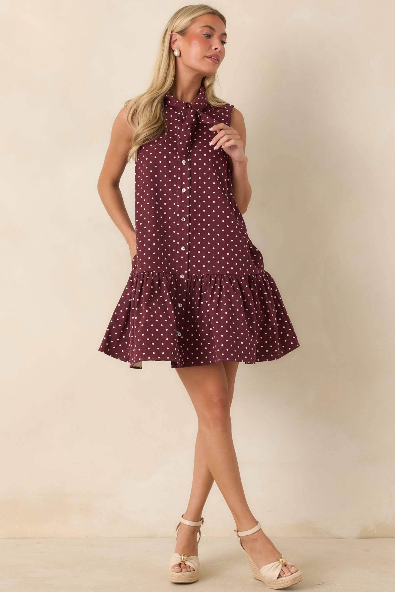 Sleeveless polka dot mini dress showcasing a self-tie neckline, button-front design, gathered ruffle hem, and side pockets.