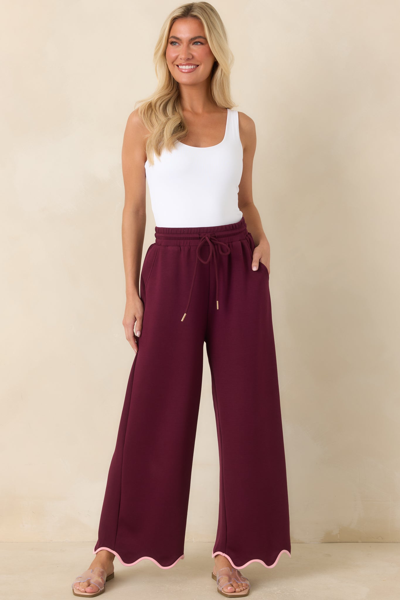 Soft burgundy pull-on pants with flowing wide legs, functional side pockets, and contrasting blush pink scalloped detailing along the hemline.