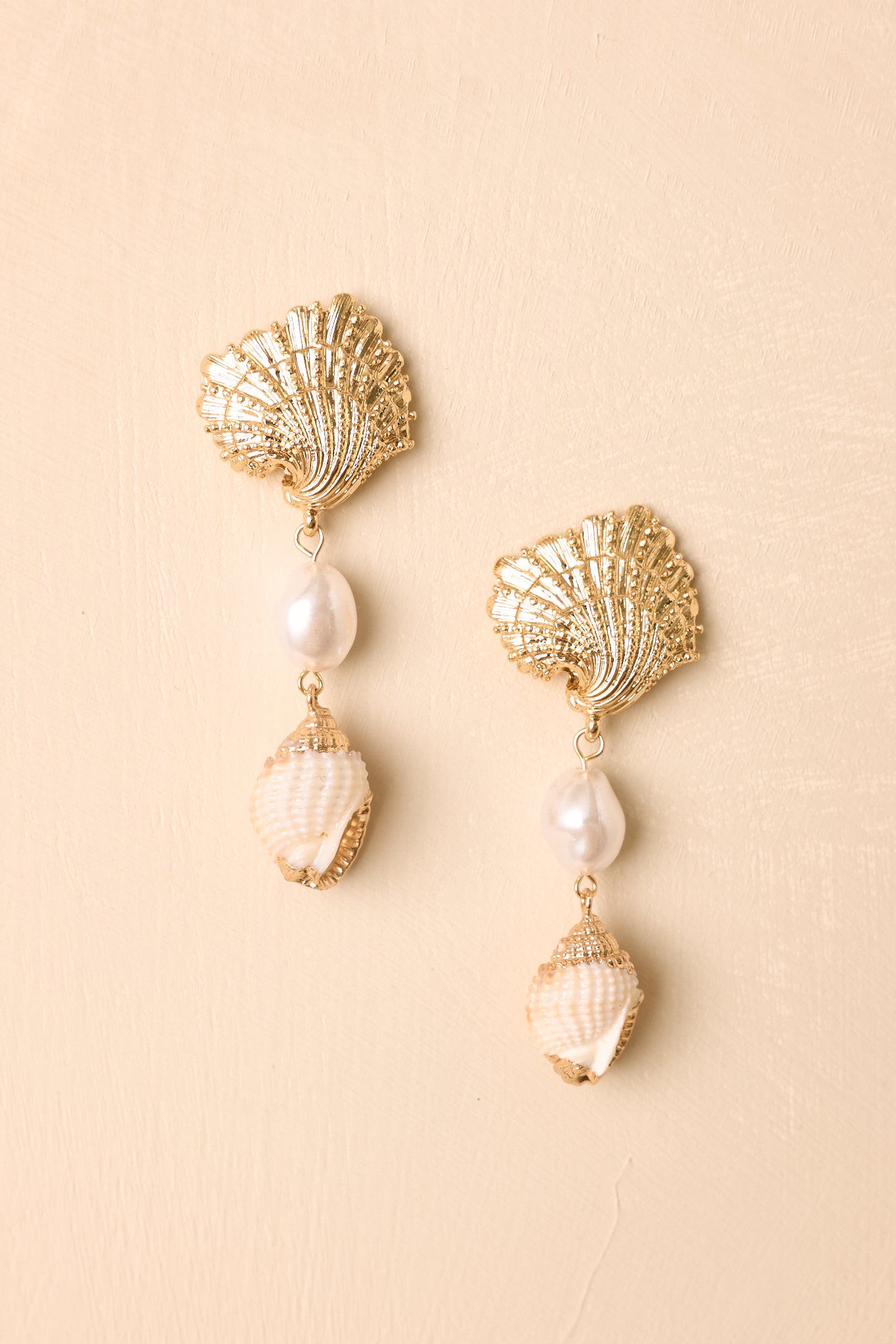 Statement earrings arranged on a neutral beige background featuring polished gold details and soft pearl accents.