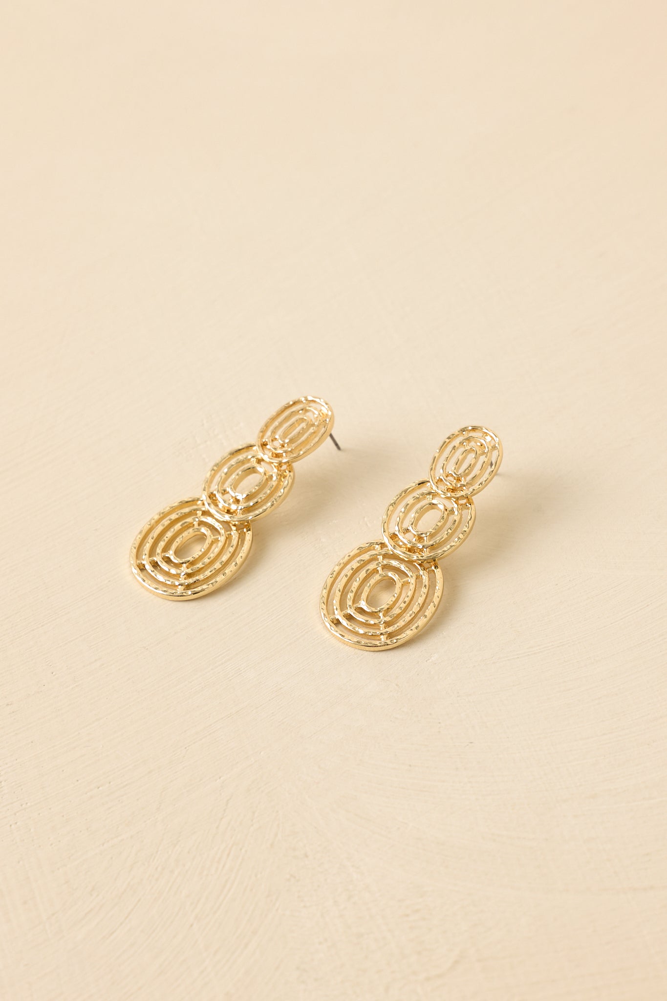 Statement gold earrings with stacked open circles and a dimensional design shown on a beige backdrop.