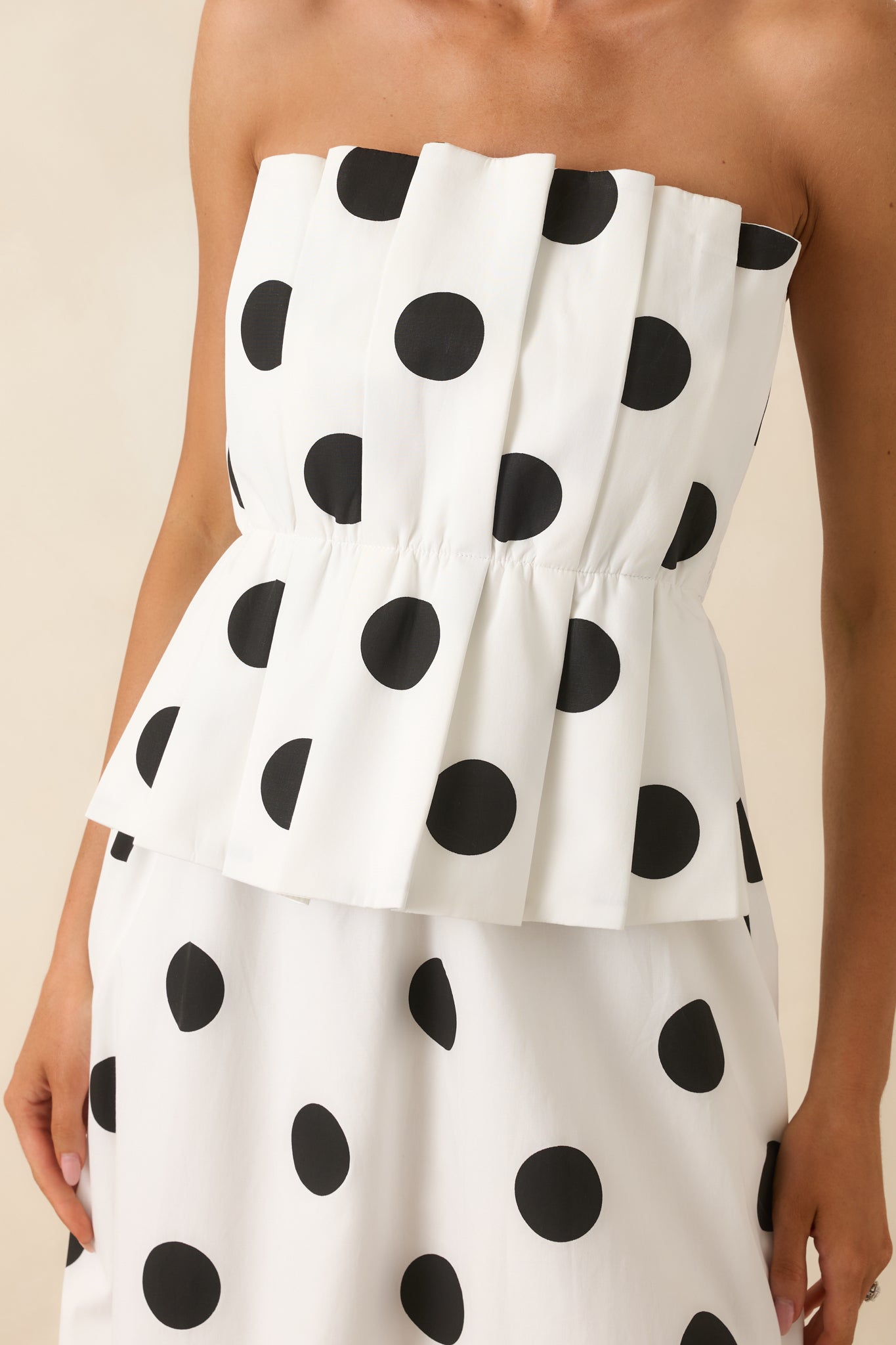 Close-up of the strapless neckline, oversized black polka dot print, structured pleated overlay, and carefully tailored construction of the midi dress.